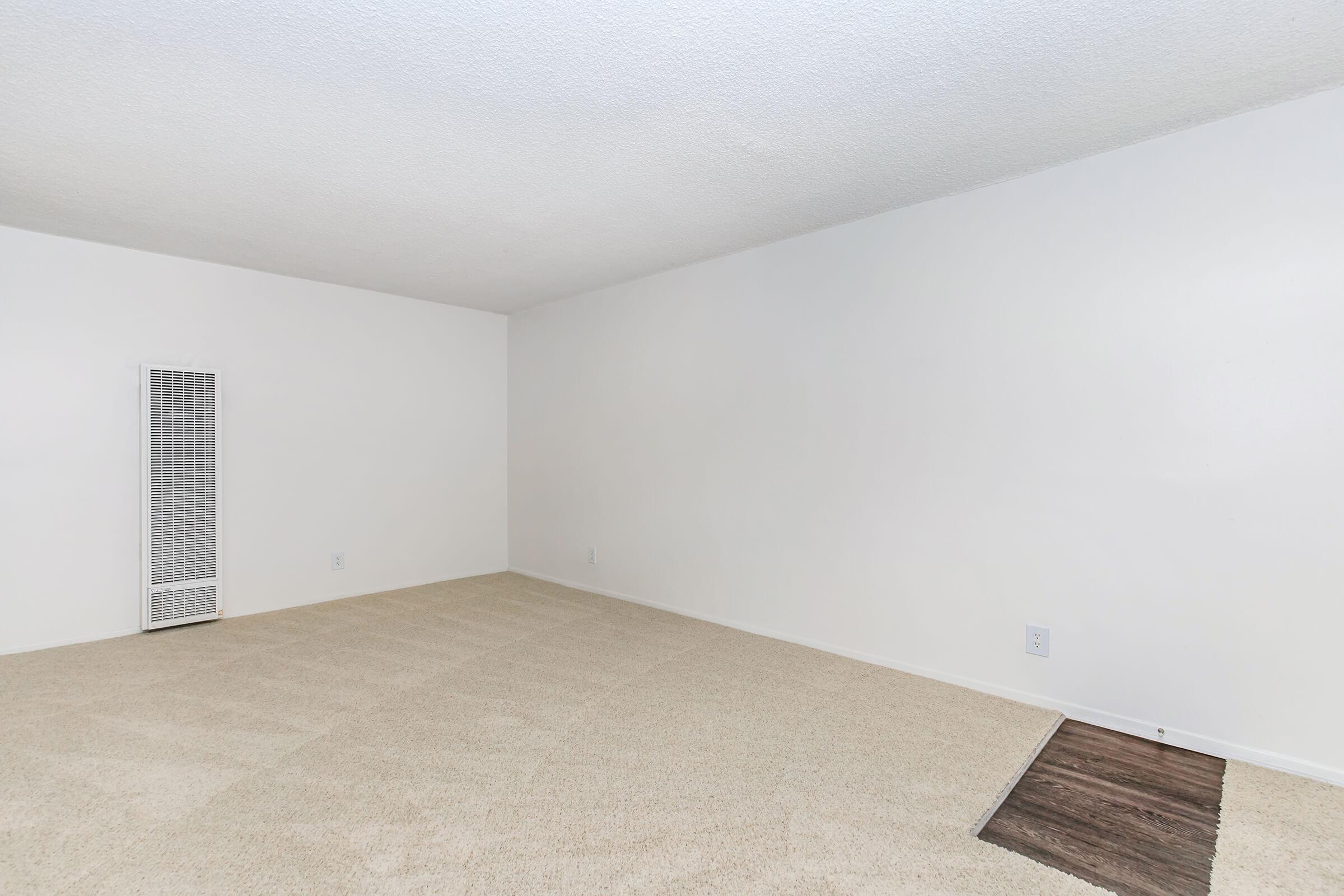 Empty room with light beige carpet and plain white walls. A small vent is visible on the left side, and a section of dark floor is exposed near the bottom right corner. The room appears spacious and well-lit, suggesting a minimalist design.