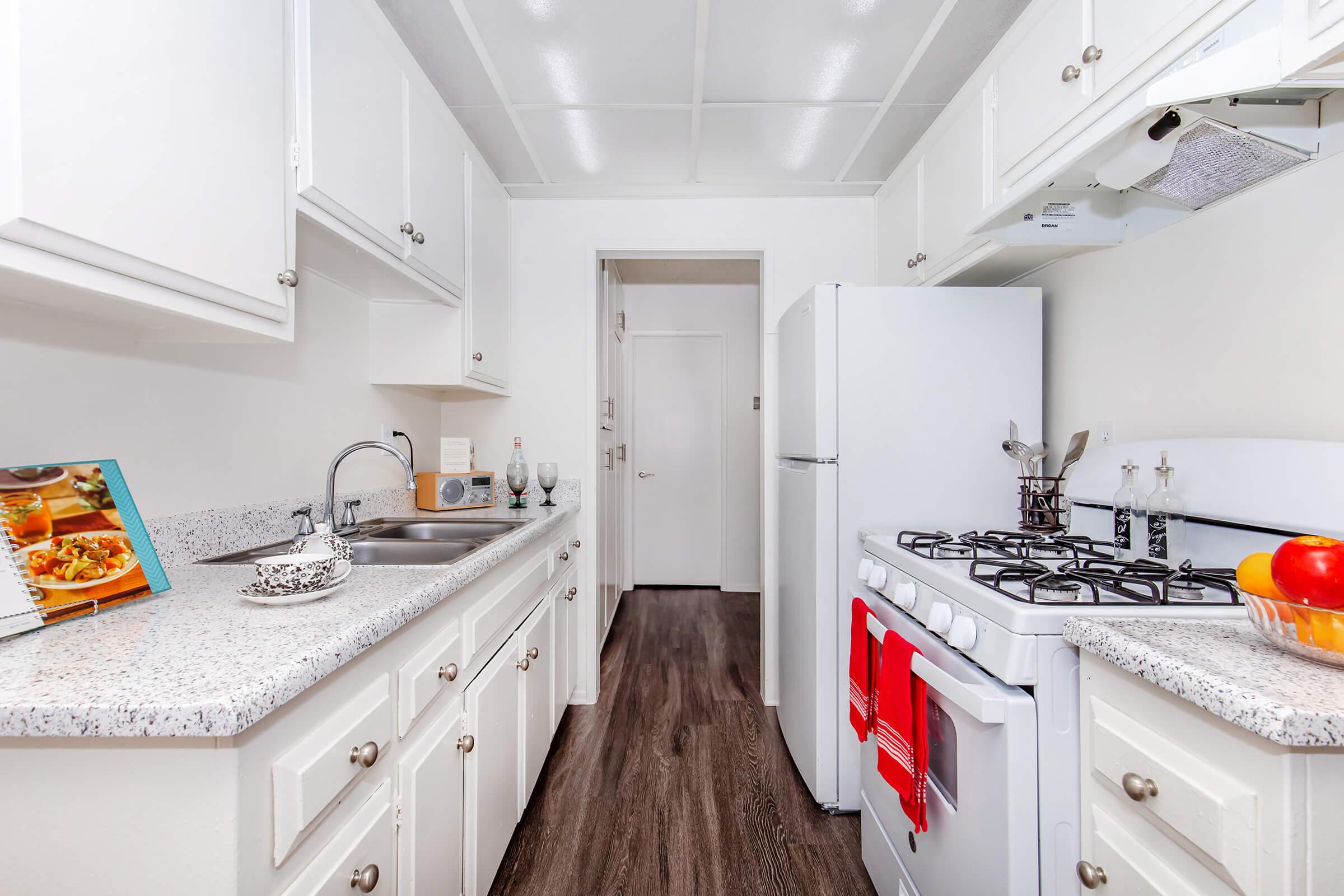 A well-lit kitchen with white cabinetry, a granite countertop, and a stainless steel sink. There’s a gas stove with a red oven mitt, a white refrigerator, and a bowl of apples on the counter. Cookbooks and decorative items are arranged on the countertop. The flooring is dark wood, adding contrast to the bright space.