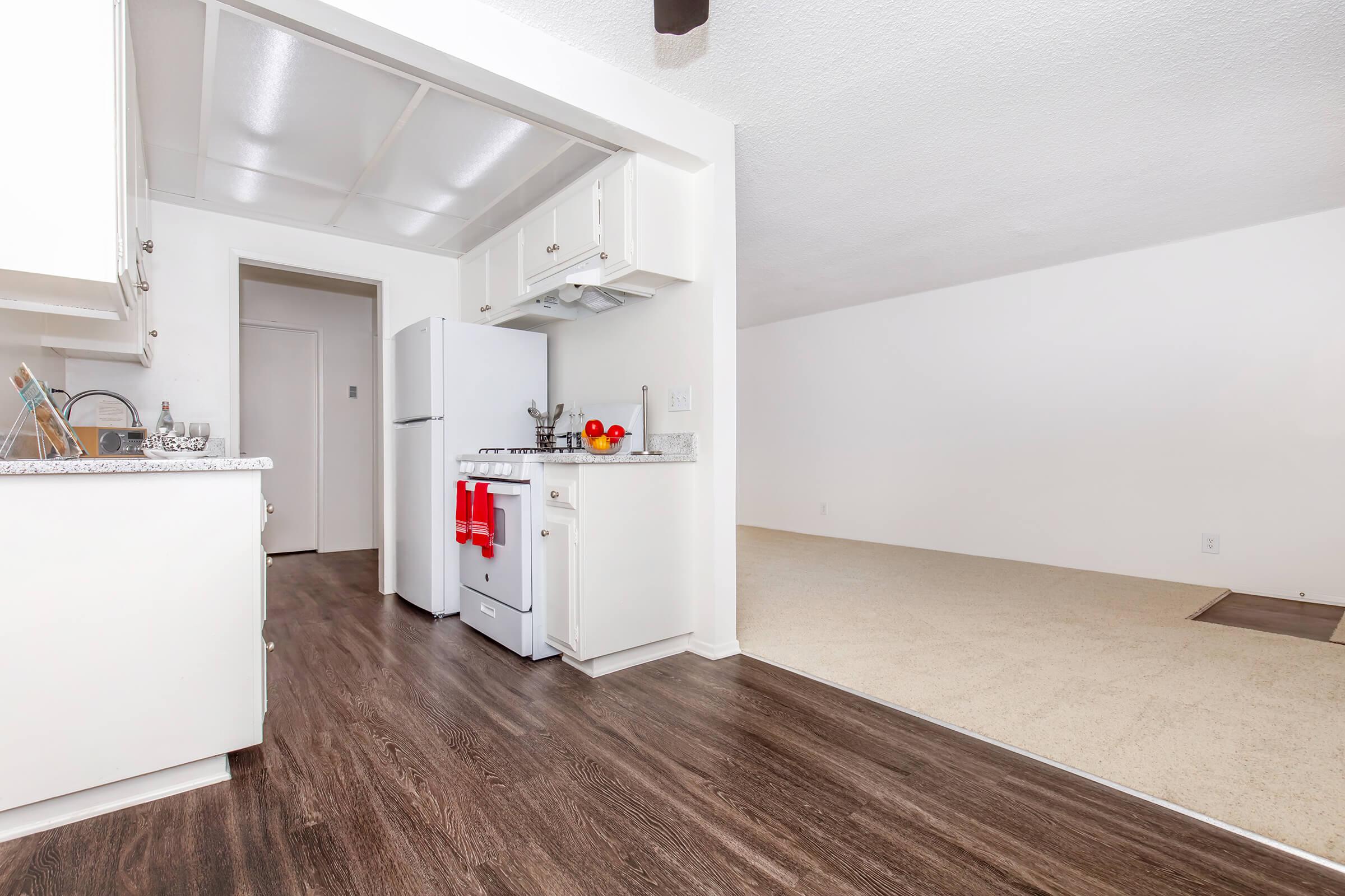 A modern kitchen featuring white cabinetry, a stove, and refrigerator. The countertop is adorned with fruit, and a red dish towel hangs on the oven. The adjacent area has light-colored carpet and white walls, creating a spacious and inviting atmosphere.