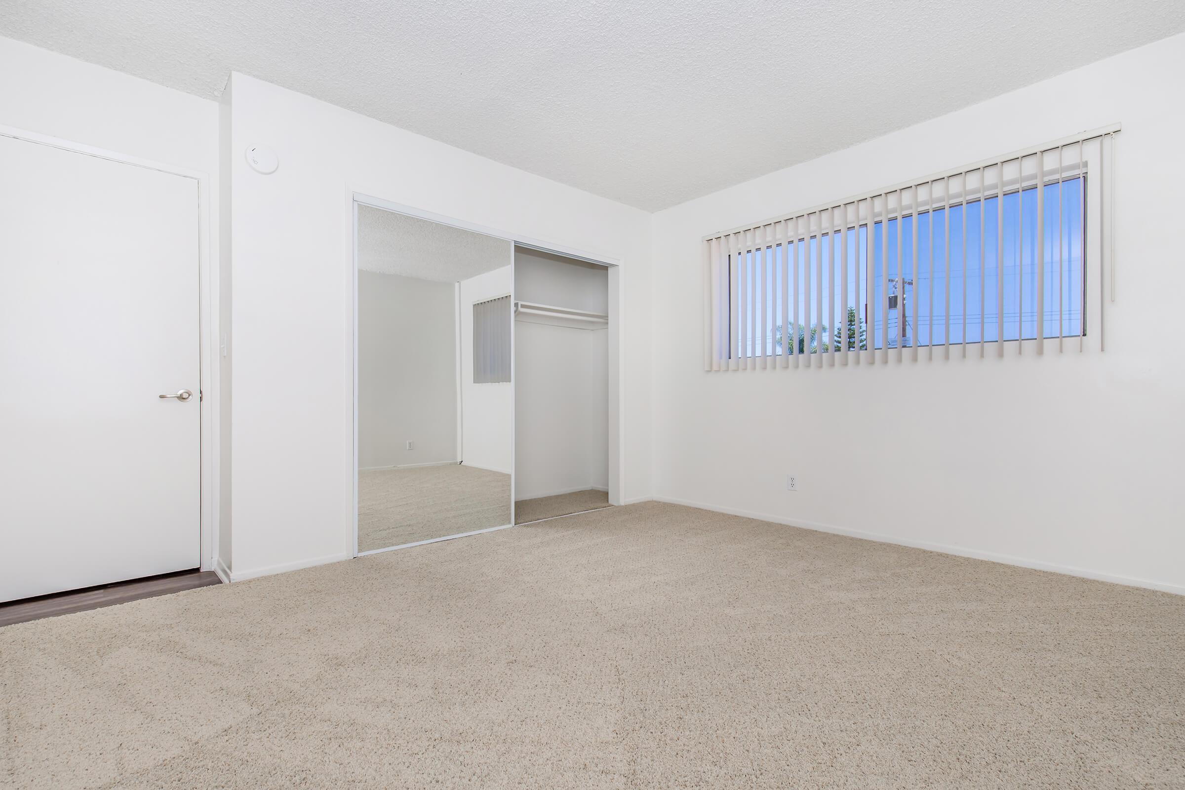 A bright, empty room with light-colored carpet and white walls. There is a closet with mirrored sliding doors on one side and a window with vertical blinds letting in natural light. The floor is clear, creating a spacious look.