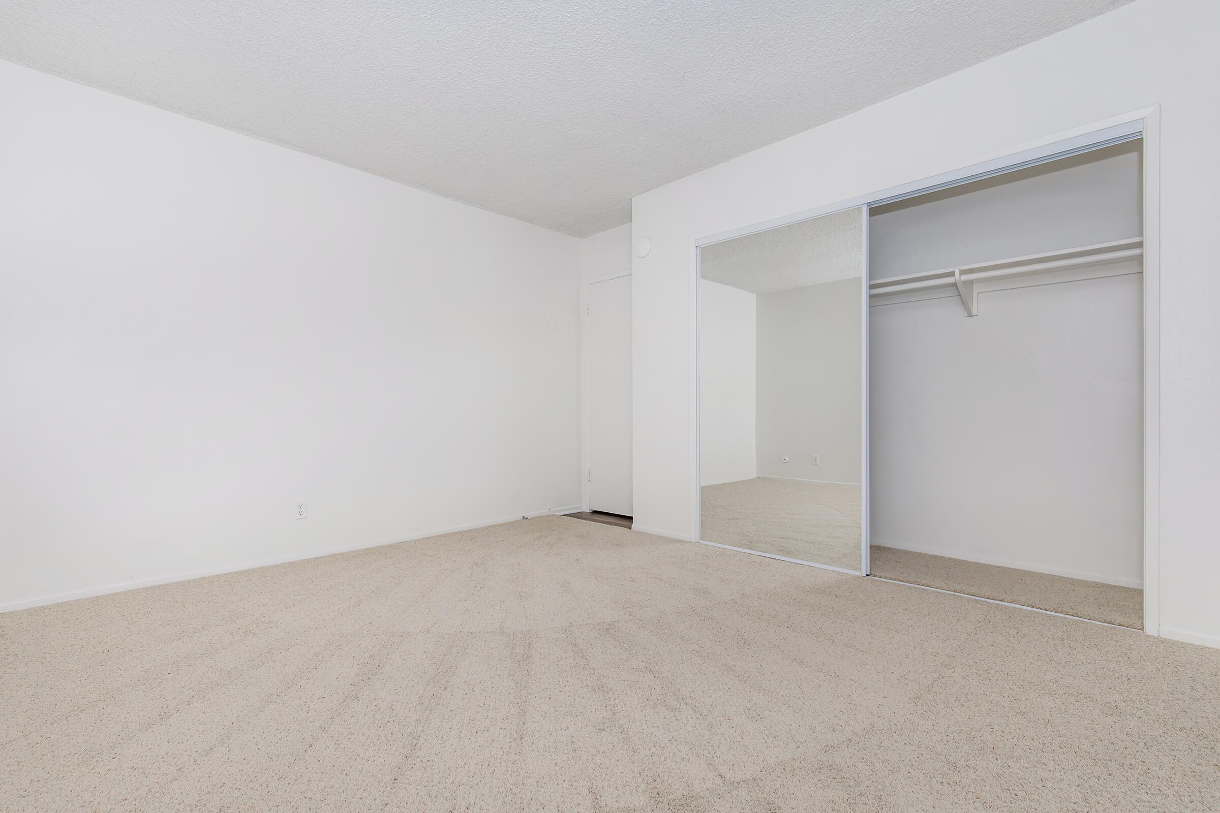 Empty room with beige carpet, white walls, and a mirrored closet on one side. Natural light fills the space, creating an airy feel. The room has no furniture, highlighting its open layout and potential for personalization.