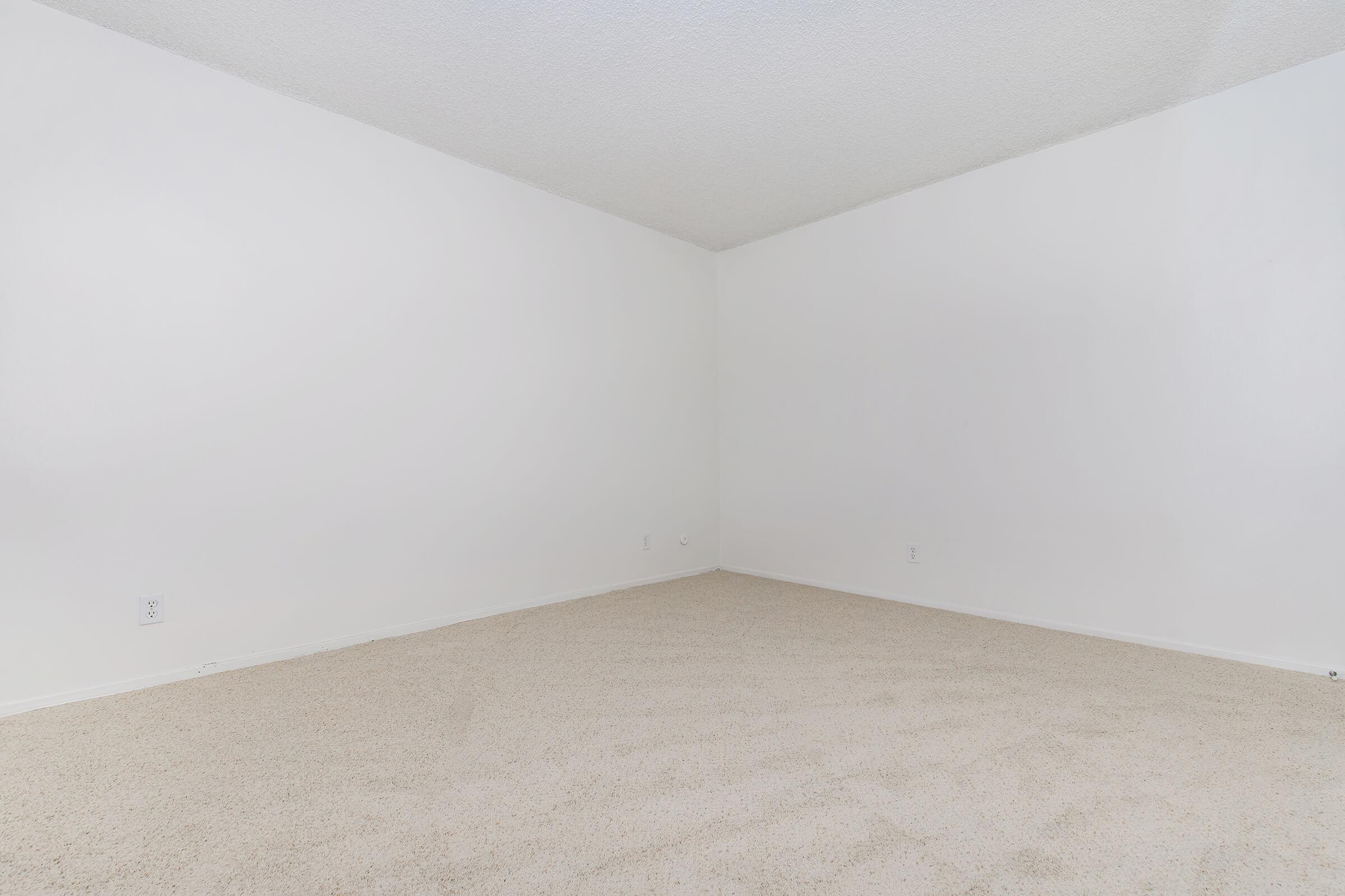 Empty room with white walls and a light beige carpet. The space is devoid of furniture or decorations, featuring two walls that meet at a right angle. Natural light may come from unseen windows, creating a bright and minimalistic atmosphere.