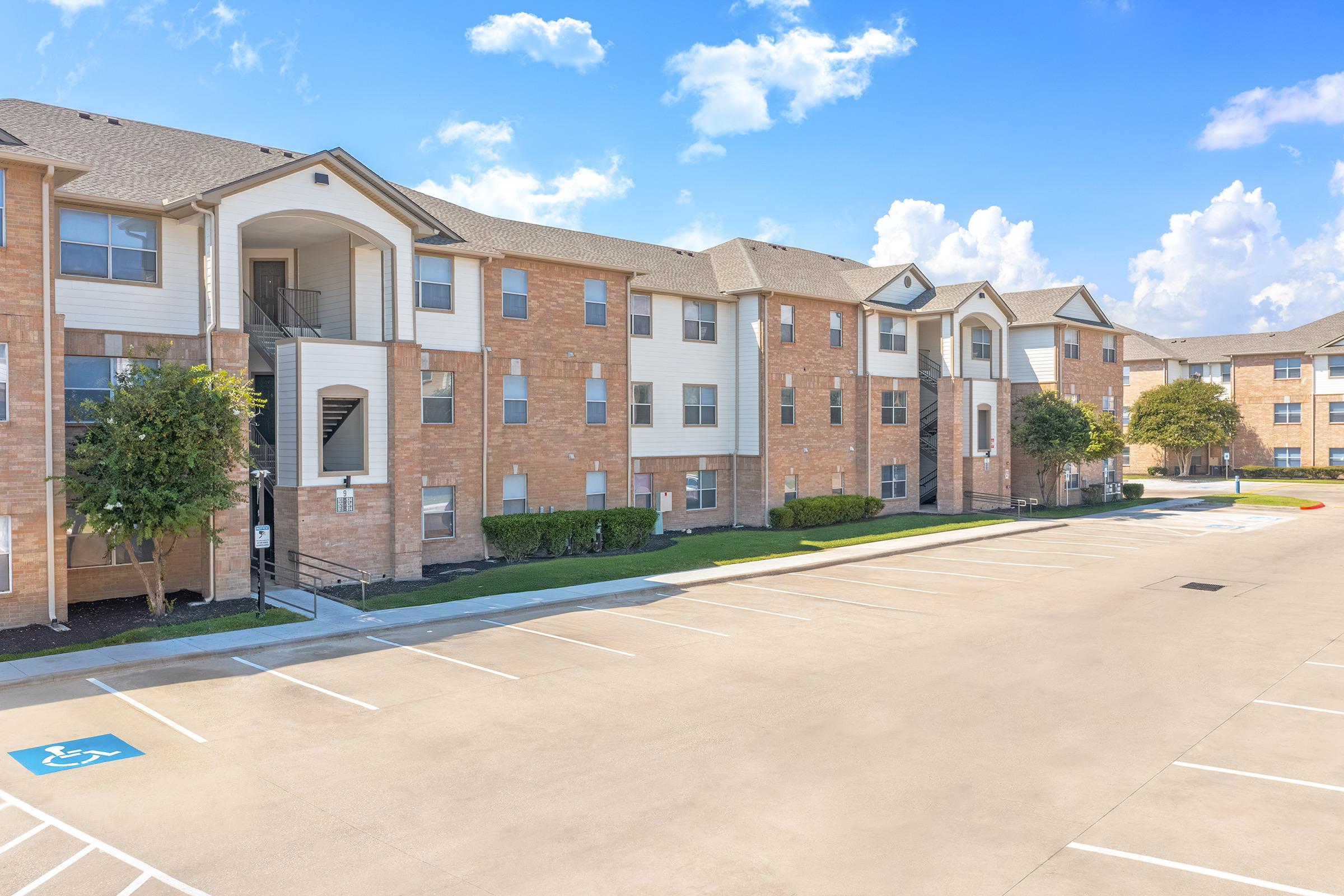 A view of a modern apartment complex featuring multiple brick buildings, well-maintained landscaping, and a spacious parking lot. The sky is blue with some clouds, creating a bright and inviting atmosphere.
