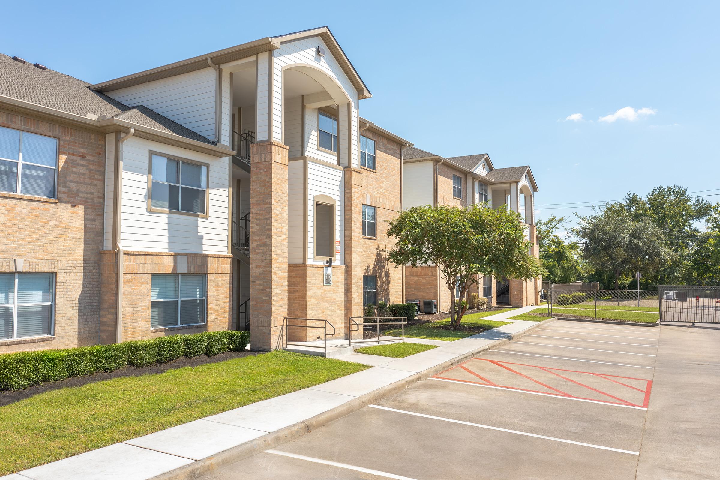 A well-maintained apartment building with brick exteriors and white accents. The structure features multiple windows, a staircase leading to the entrance, and landscaping with green grass and small shrubs. A parking lot is visible in front, with designated parking spaces. The sky is clear and blue.