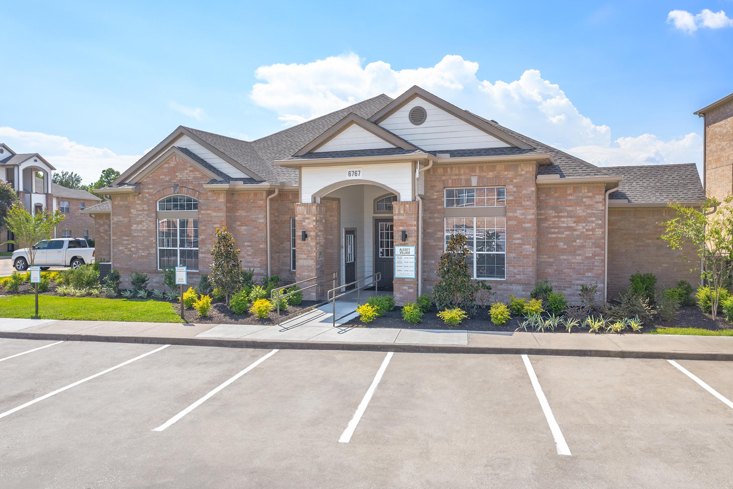 A modern brick building with large windows, featuring a central entrance and surrounded by landscaped greenery. The sun is shining under a clear blue sky, and there are a few parking spaces in front, with one occupied by a vehicle. The overall setting appears welcoming and well-maintained.