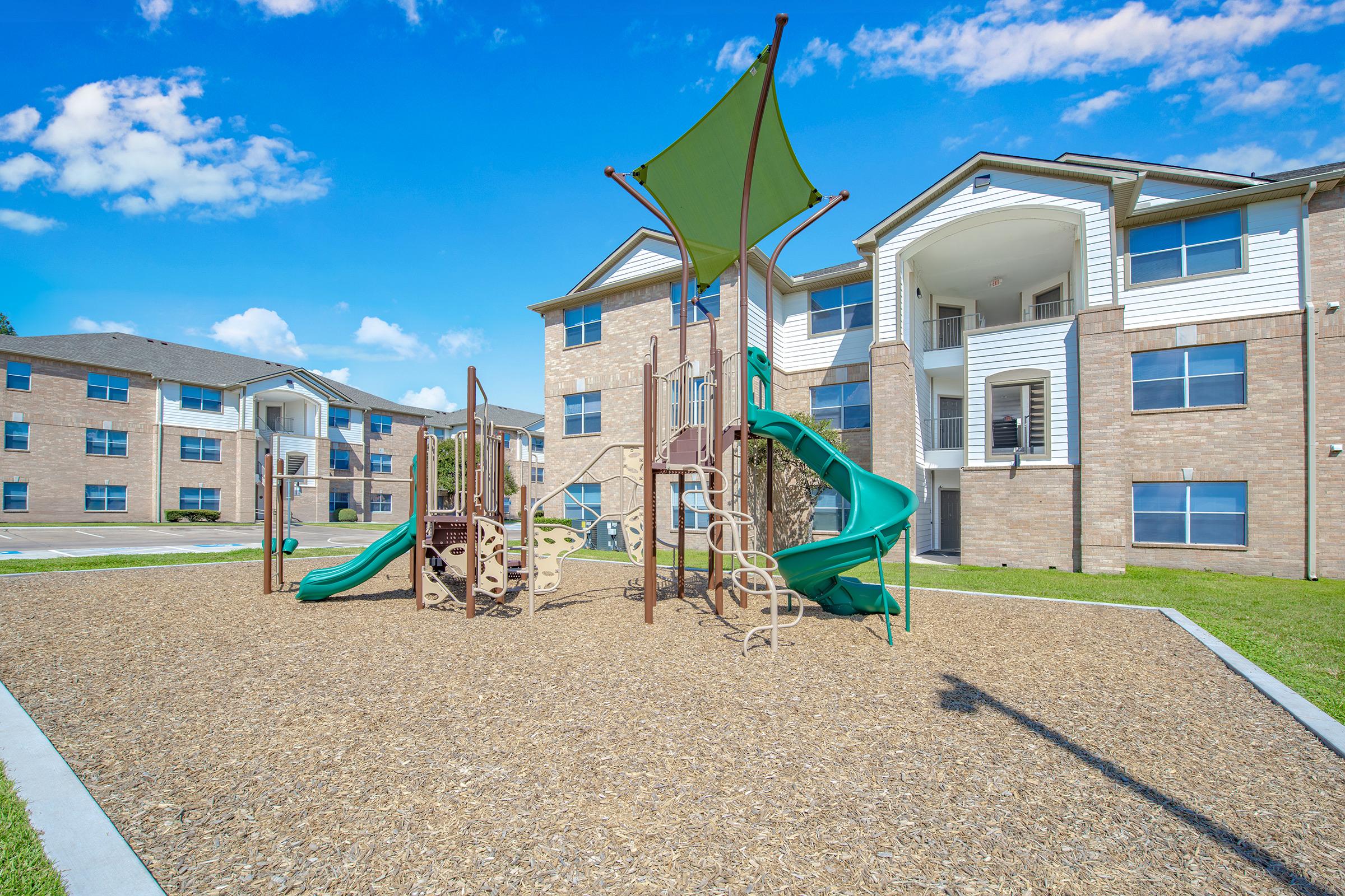 Playground area featuring green slides, climbing structures, and a shaded canopy, situated on a mulch-covered ground outside a brick apartment complex under a blue sky with scattered clouds.