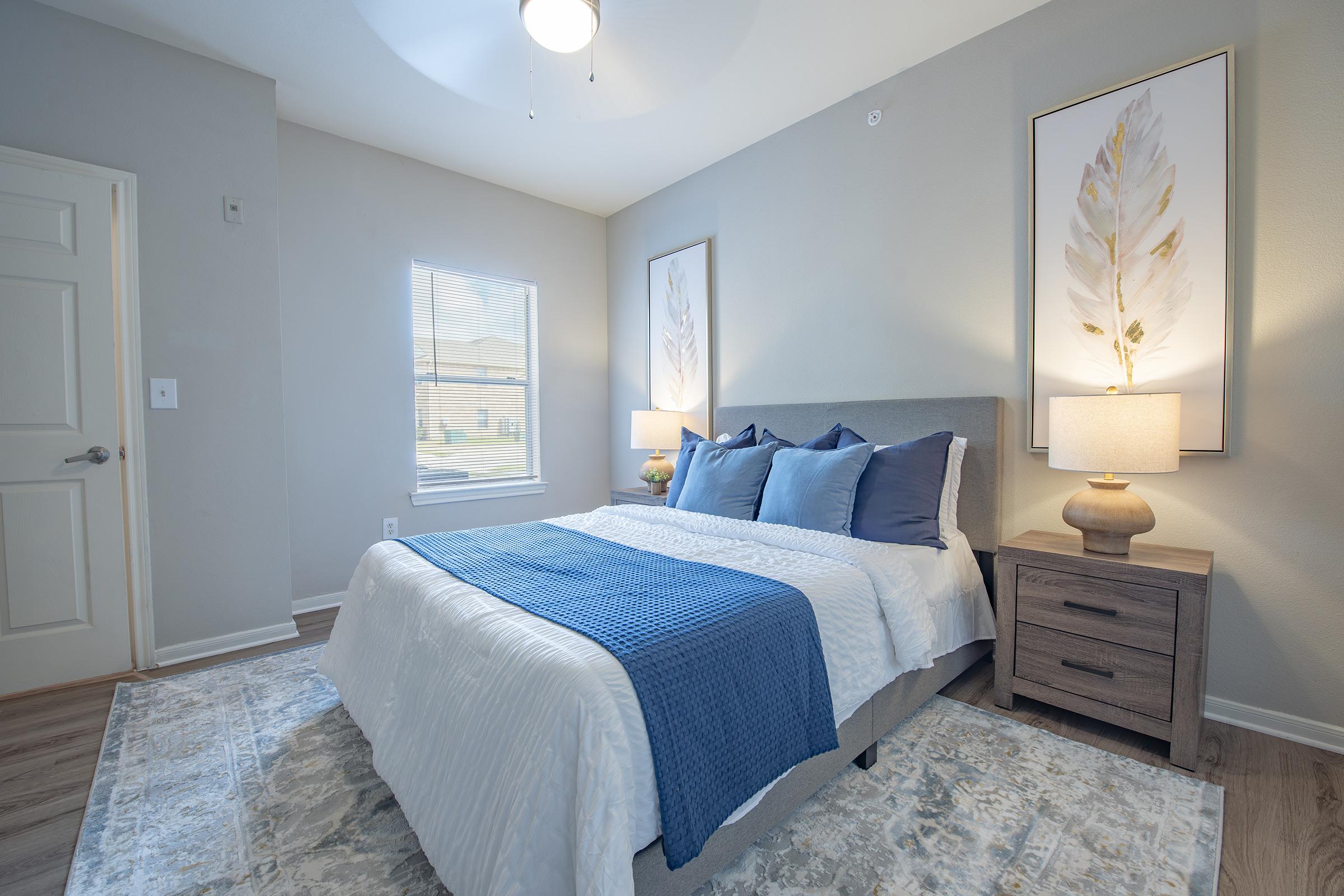 A cozy bedroom featuring a queen-sized bed with blue and white bedding, two decorative pillows, and a light gray headboard. There are two stylish lamps on nightstands flanking the bed, and two framed botanical prints on the walls. Natural light streams in through a window, illuminating the soft neutral tones of the decor.