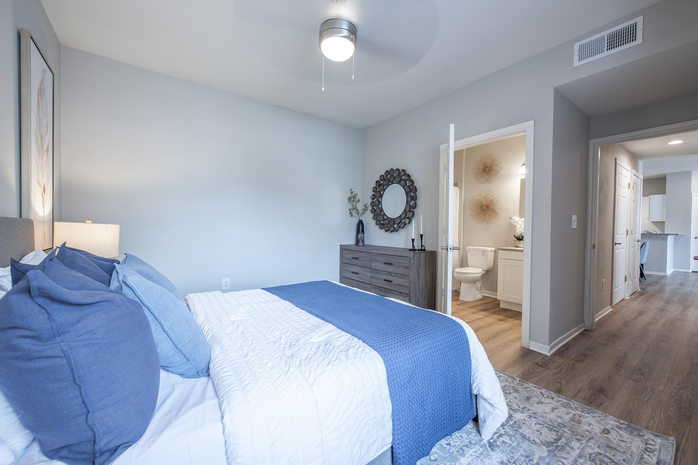 A cozy bedroom featuring a bed with blue and white bedding, a nightstand with lamps, and a decorative mirror above a dresser. A doorway leads to a bathroom with light-colored walls and modern fixtures. Natural light enhances the serene atmosphere of the space.