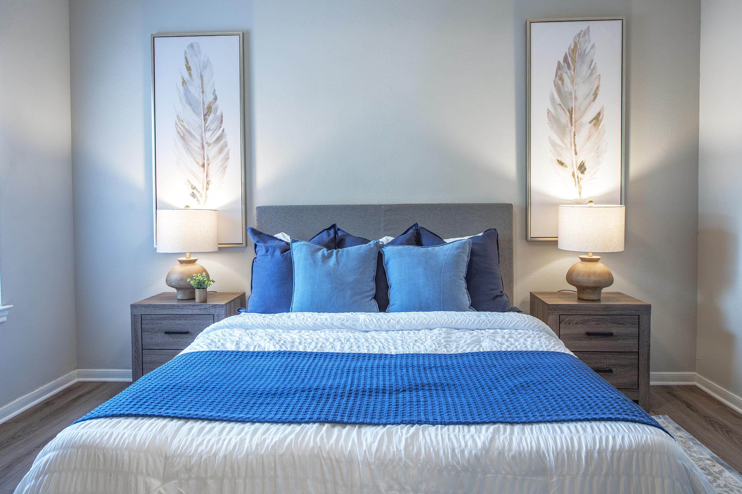 A cozy bedroom featuring a gray upholstered bed with blue and white bedding. Two stylish lamps on wooden nightstands flank the bed, and above the nightstands are framed artworks of feathers. The walls are painted light, creating a serene atmosphere with warm wooden flooring.