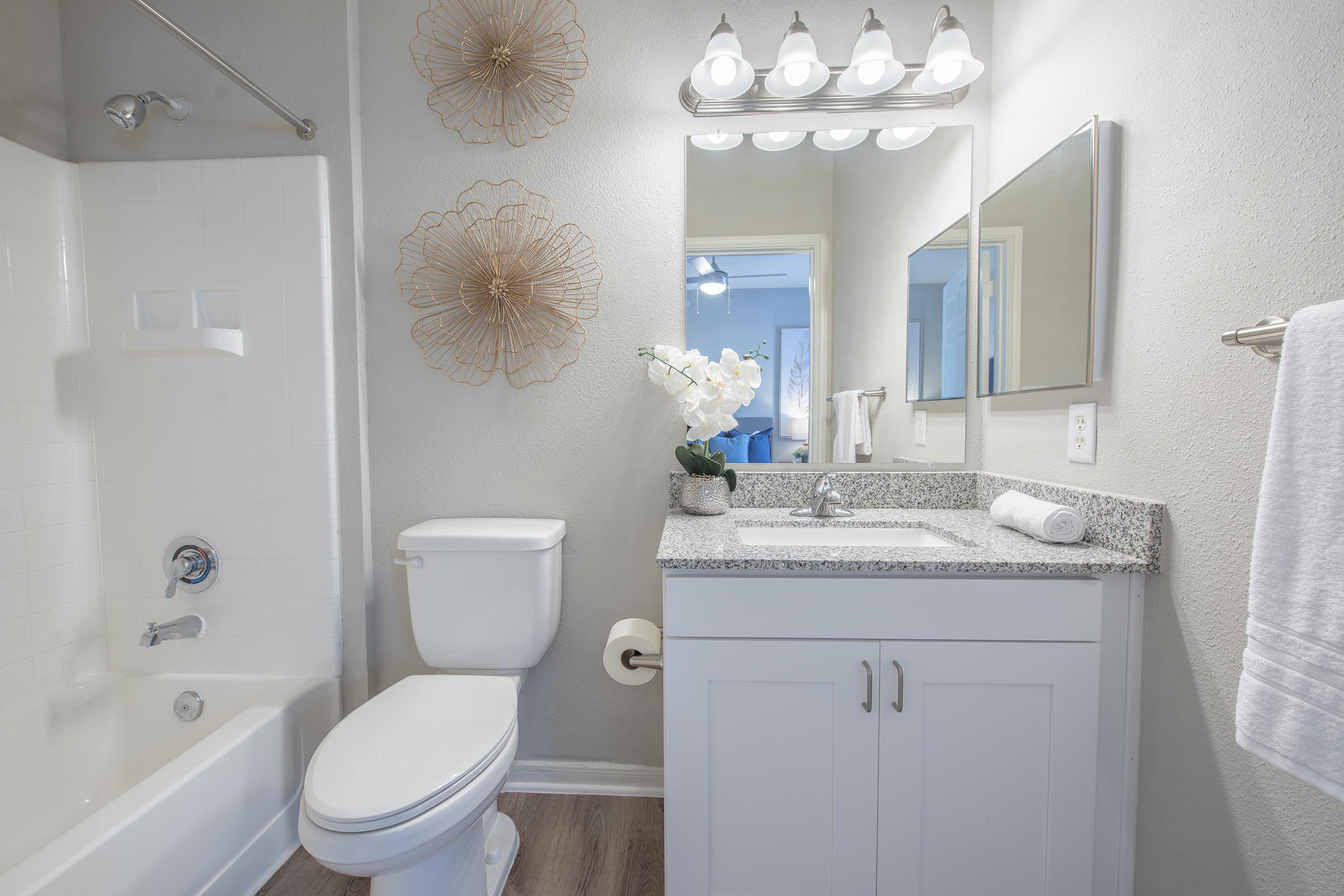 A modern bathroom featuring a white bathtub, a toilet, and a granite countertop with a sink. Above the mirror, there are stylish light fixtures. Decorative wall accents include two floral wall hangings. A potted orchid adds a touch of greenery, and a hand towel is neatly hung on the side.