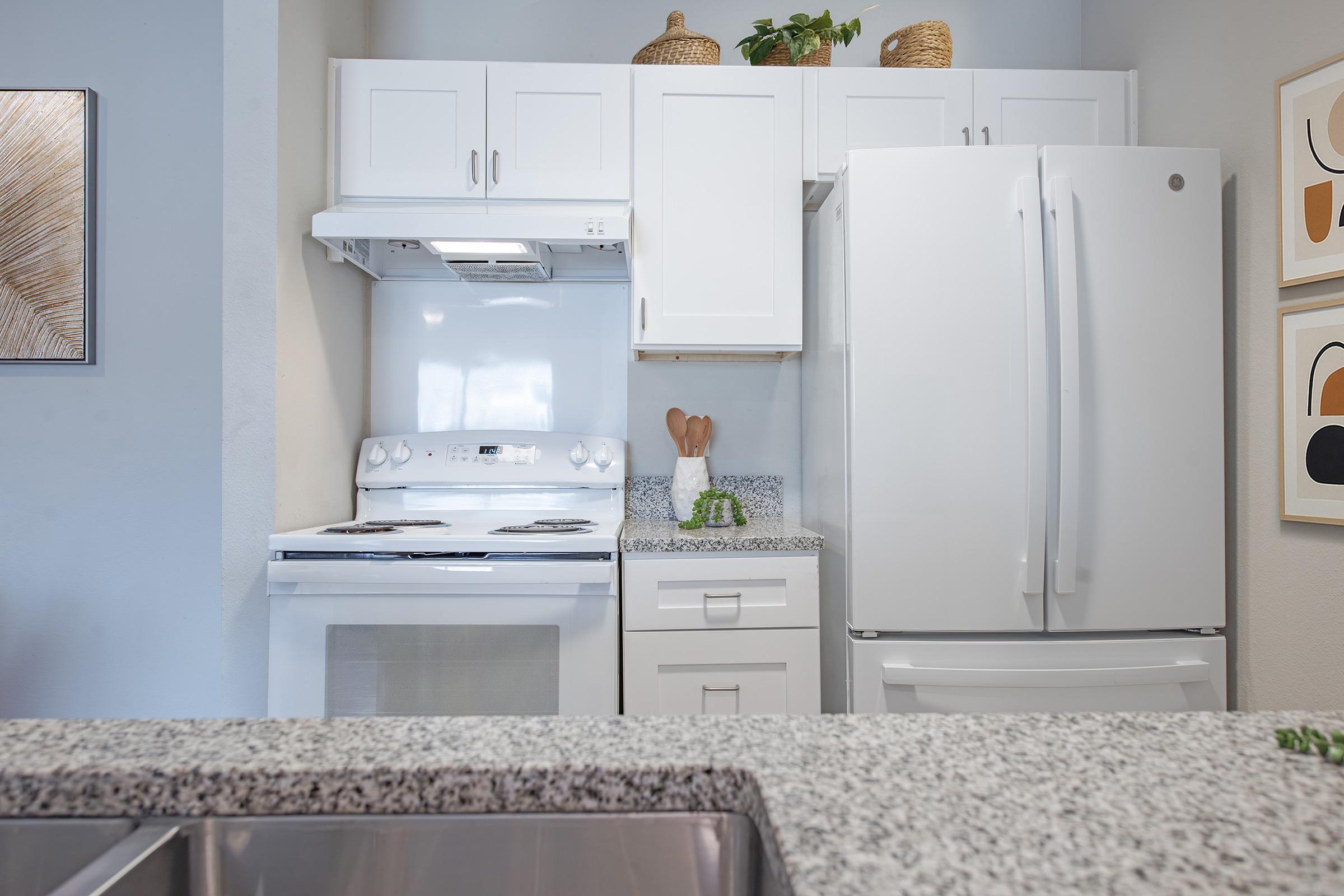 A modern kitchen featuring white cabinetry, a granite countertop, a white refrigerator, and a stovetop. The design includes decorative items like a plant and kitchen utensils. The area is well-lit and organized, with artwork on the walls and a clean, minimalist aesthetic.