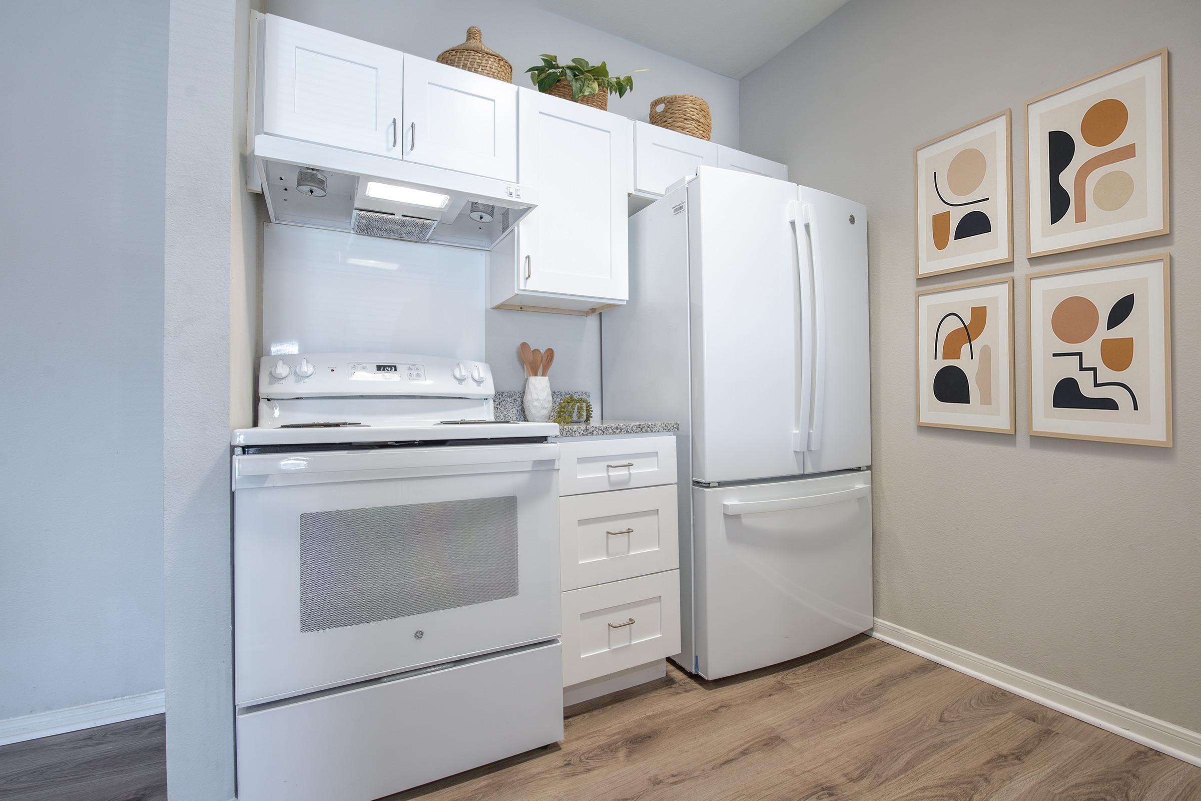 Modern kitchen featuring a white stove, ventilation hood, and refrigerator. Light cabinetry complements the clean design. On the wall, there are four abstract framed prints with warm colors. The flooring is wooden, adding warmth to the space, while decorative items are placed on the countertop and shelves.