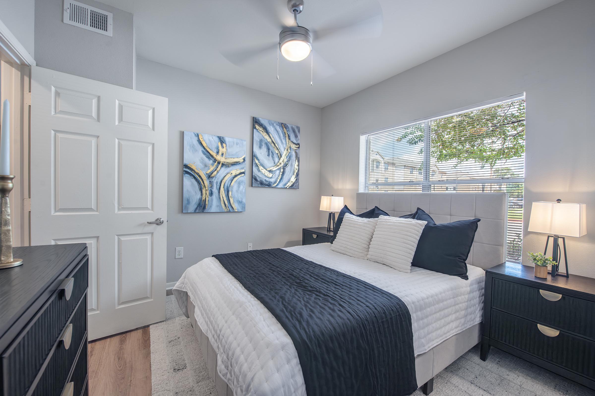 A cozy bedroom featuring a queen-sized bed with a dark blue comforter, adorned with decorative pillows. There are two abstract paintings on the wall, stylish bedside lamps on wooden nightstands, and a large window letting in natural light. A ceiling fan adds a modern touch to the room.