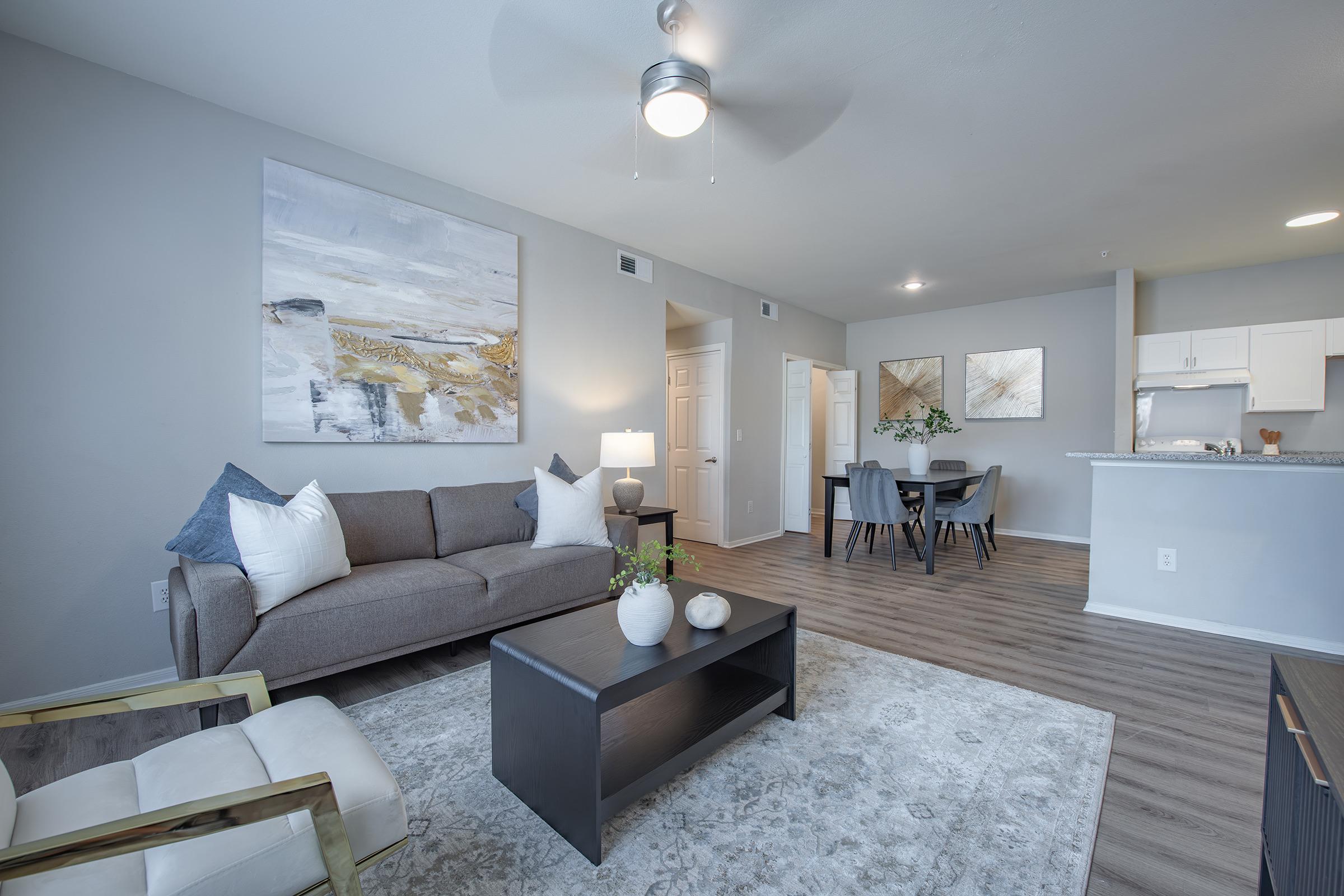 A modern living space featuring a beige sofa with blue and white pillows, a dark coffee table, and a decorative rug. A dining area with a small table is visible in the background. The walls are painted light gray, and there is a large abstract painting above the sofa. A ceiling fan provides additional lighting.