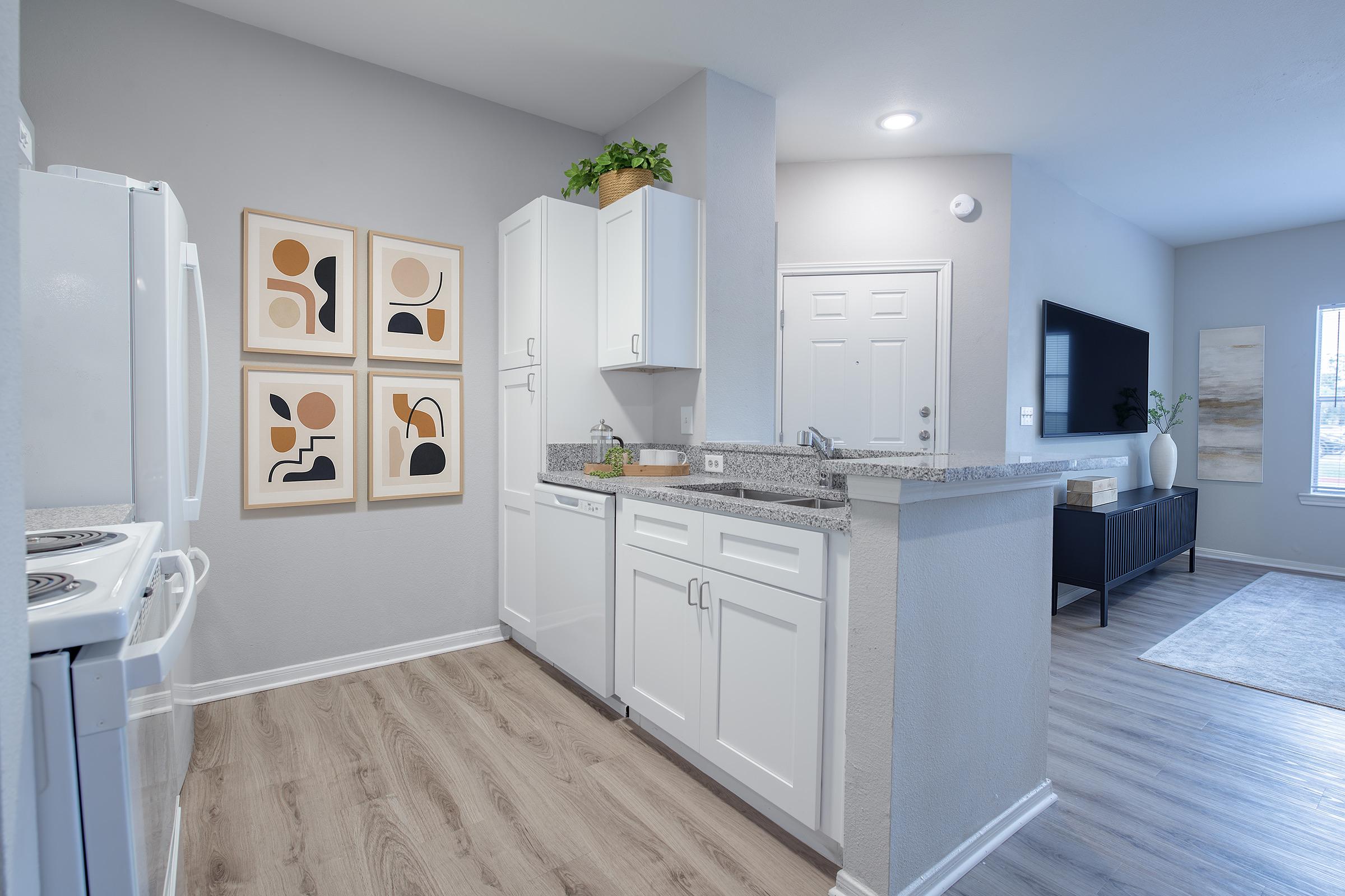 A modern kitchen with white cabinets, granite countertops, and a white refrigerator. Four abstract art pieces are hung on the wall. The space opens to a living area with a TV and a black console, featuring a large window with natural light and light wood flooring throughout.