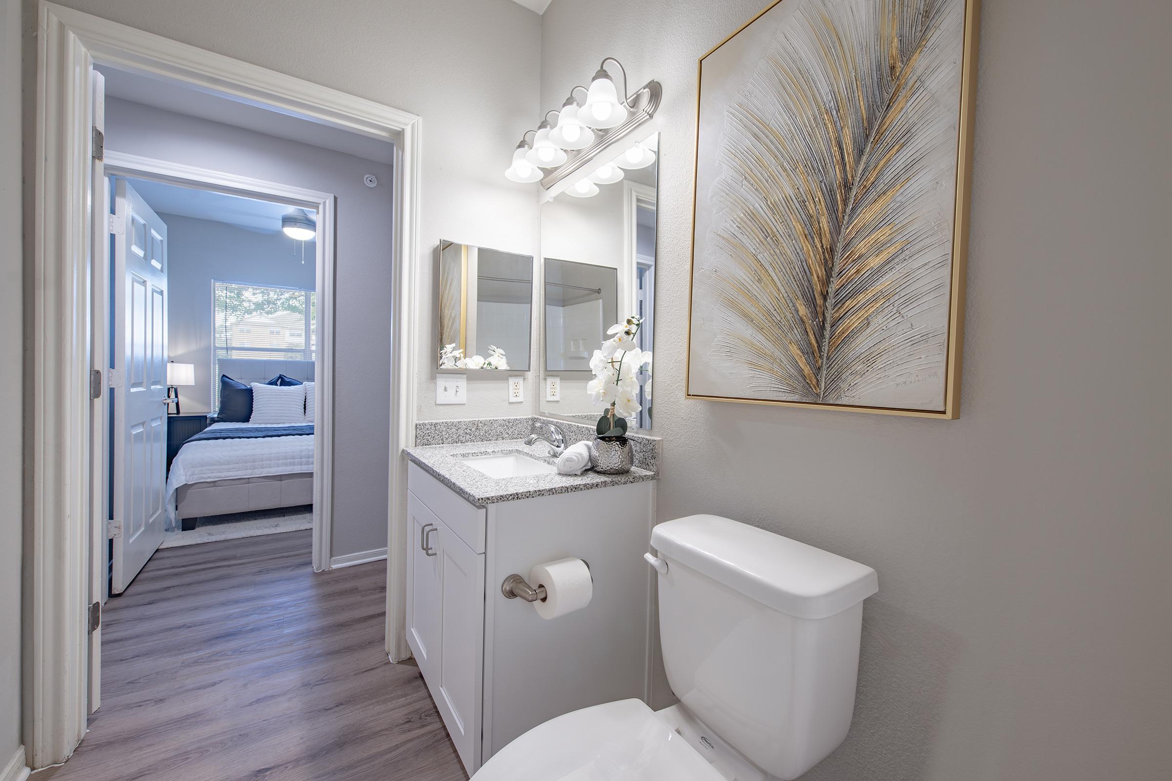 Bright bathroom with a modern design, featuring a vanity with a sink, large mirror, and decorative artwork. A white toilet is located nearby. A doorway leads to a softly lit bedroom with a bed and window visible in the background. Natural light enhances the inviting atmosphere.