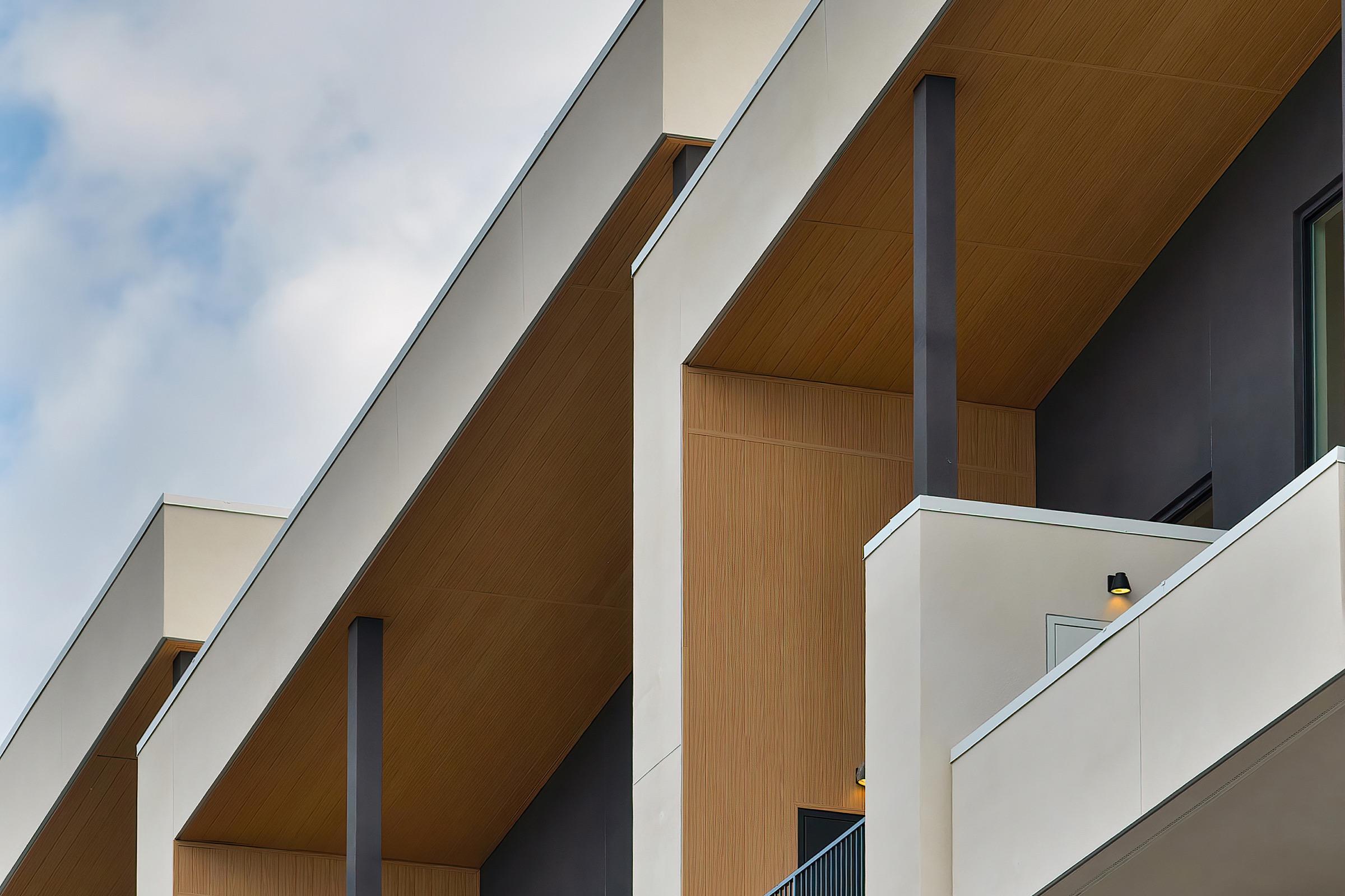 Close-up view of modern architectural design featuring multiple balconies with wooden accents and white walls. The triangular rooflines create a dynamic look against a cloudy sky, showcasing a blend of materials and contemporary style.