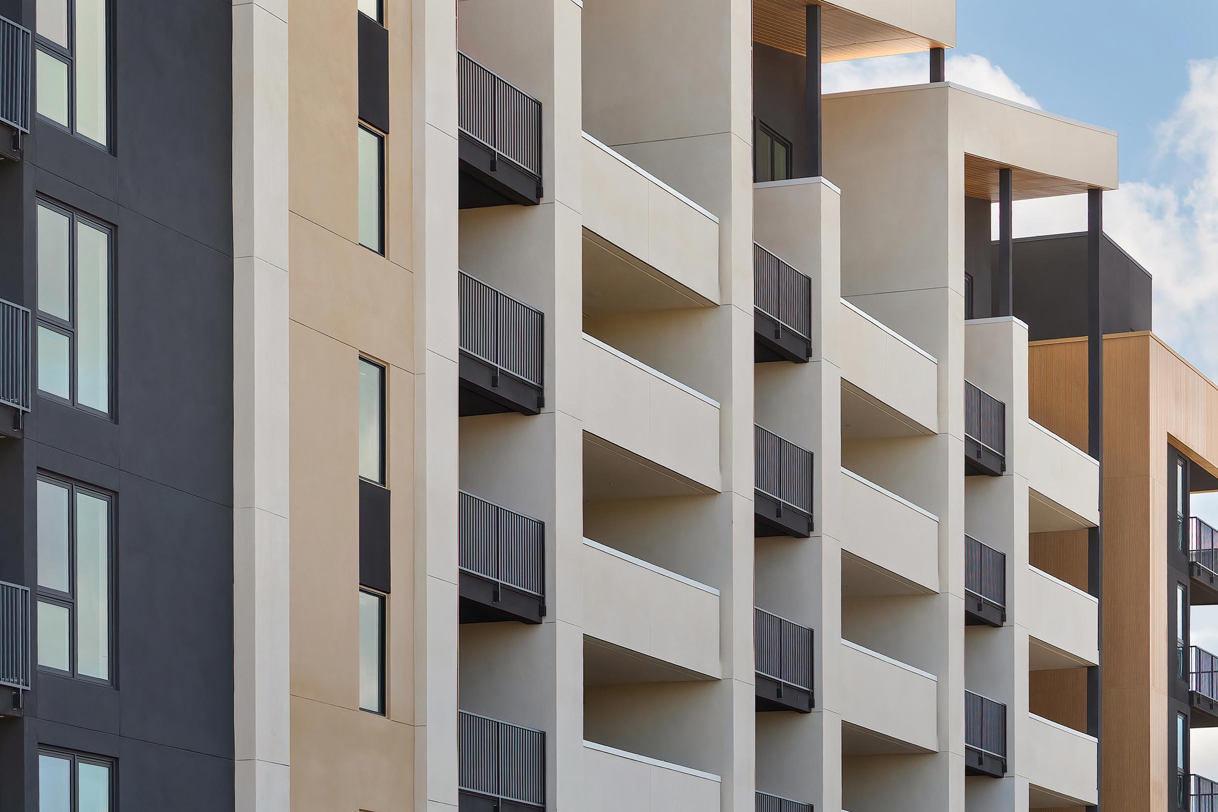 Modern apartment building facade with a mix of light-colored and dark-colored materials. Features multiple balconies with railing. The design has a contemporary aesthetic, showcasing clean lines and large windows. Soft blue sky with a few clouds in the background.