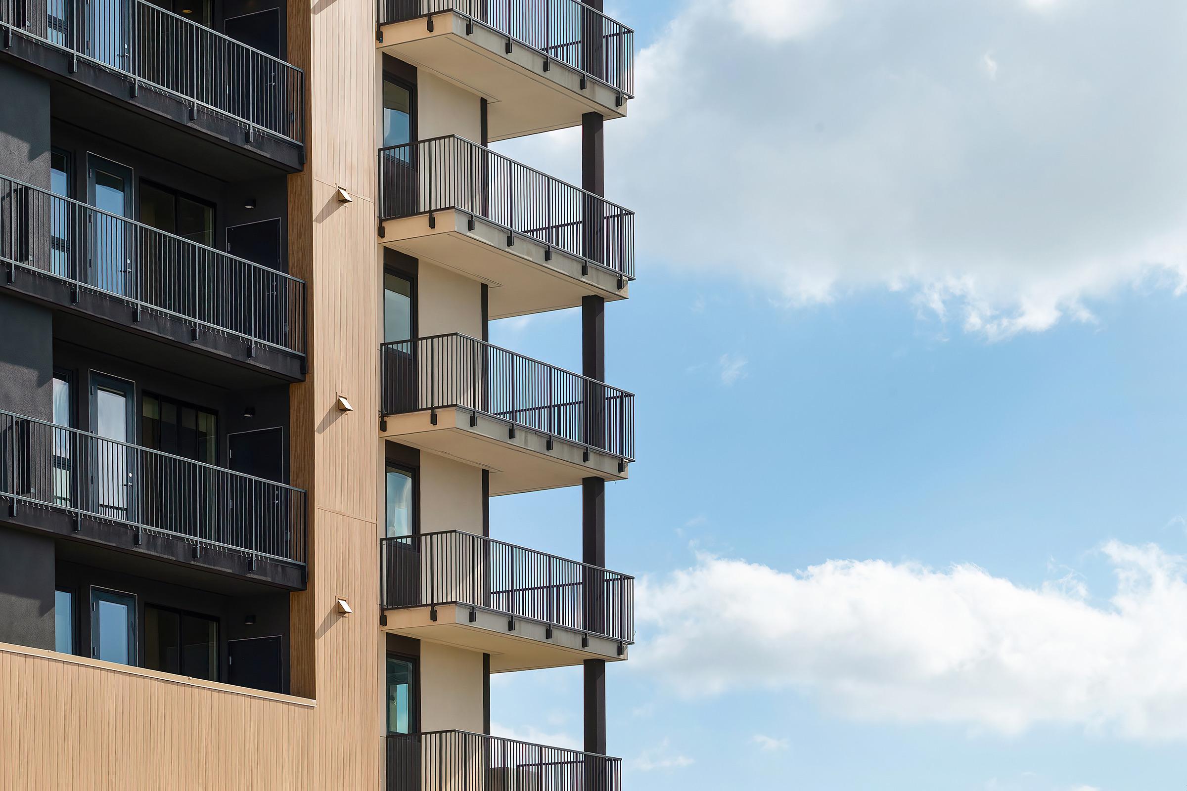 A modern building corner featuring multiple balconies with glass railings. The facade has a combination of dark and light materials, with a clear blue sky and fluffy clouds in the background. The structure exemplifies contemporary architectural design.