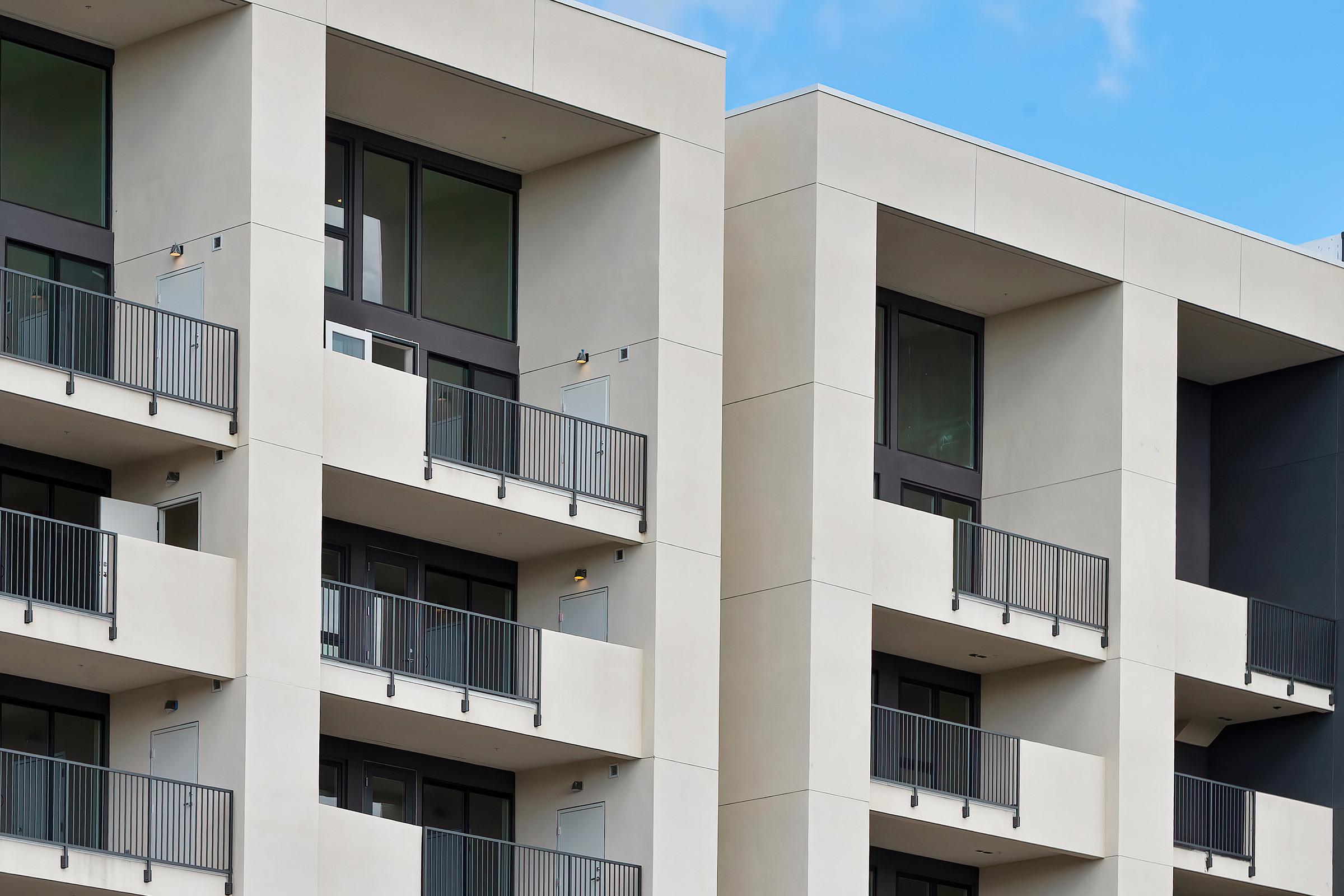 Close-up view of modern apartment building exteriors featuring sleek, understated architecture. The structure has multiple levels with balconies and large windows, showcasing a minimalist design against a blue sky.