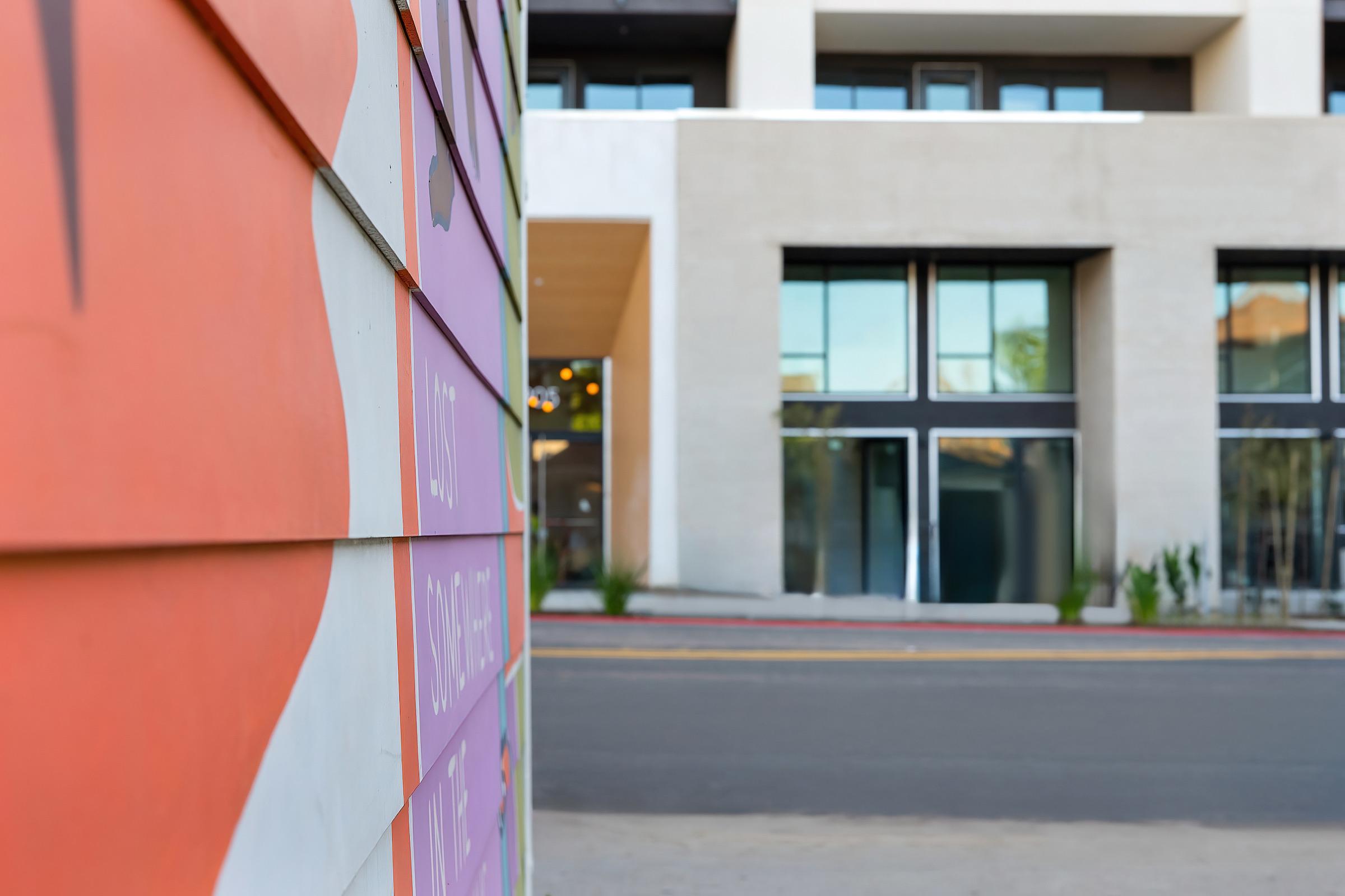 Colorful wooden panels in the foreground with text and designs, featuring vibrant hues of orange and purple. In the background, a modern building with large windows and a minimalist design, set against a clear sky. The scene captures a blend of artistic elements and contemporary architecture.