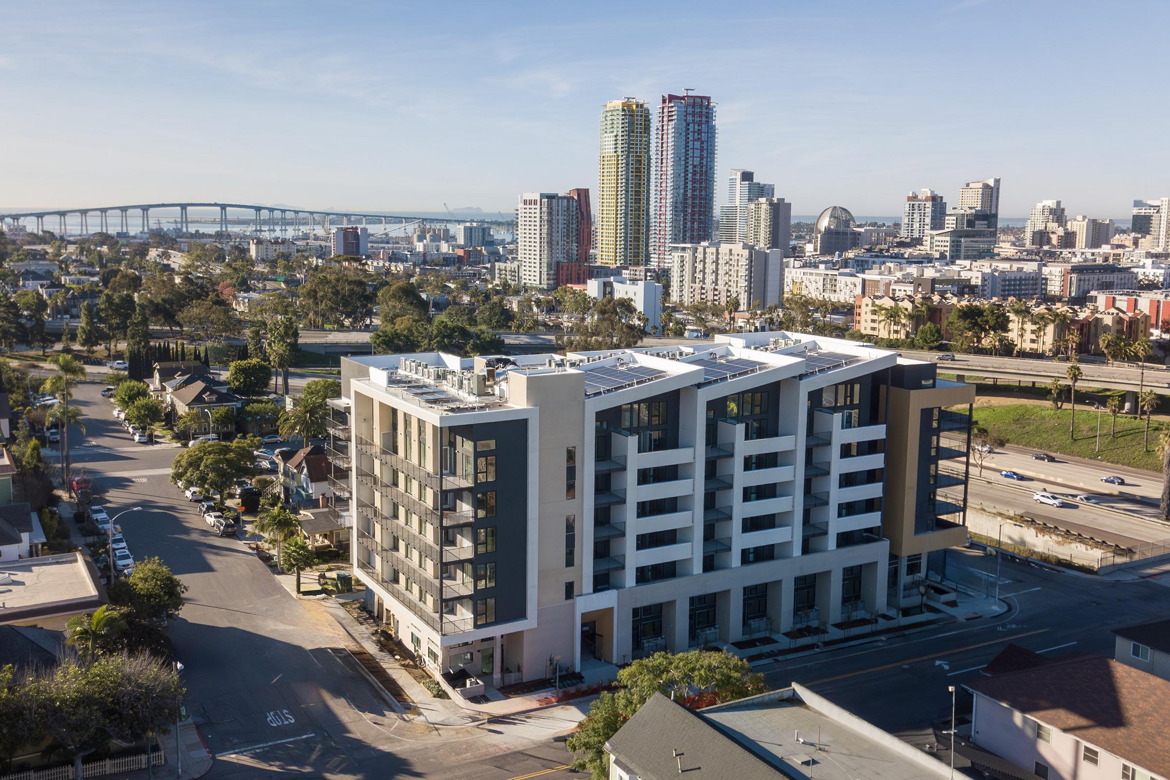 Aerial view of a modern multi-story apartment building in the foreground, surrounded by streets and trees. In the background, a skyline featuring tall buildings and a bridge under a clear blue sky. The scene captures urban architecture and city life with well-defined structures and greenery.