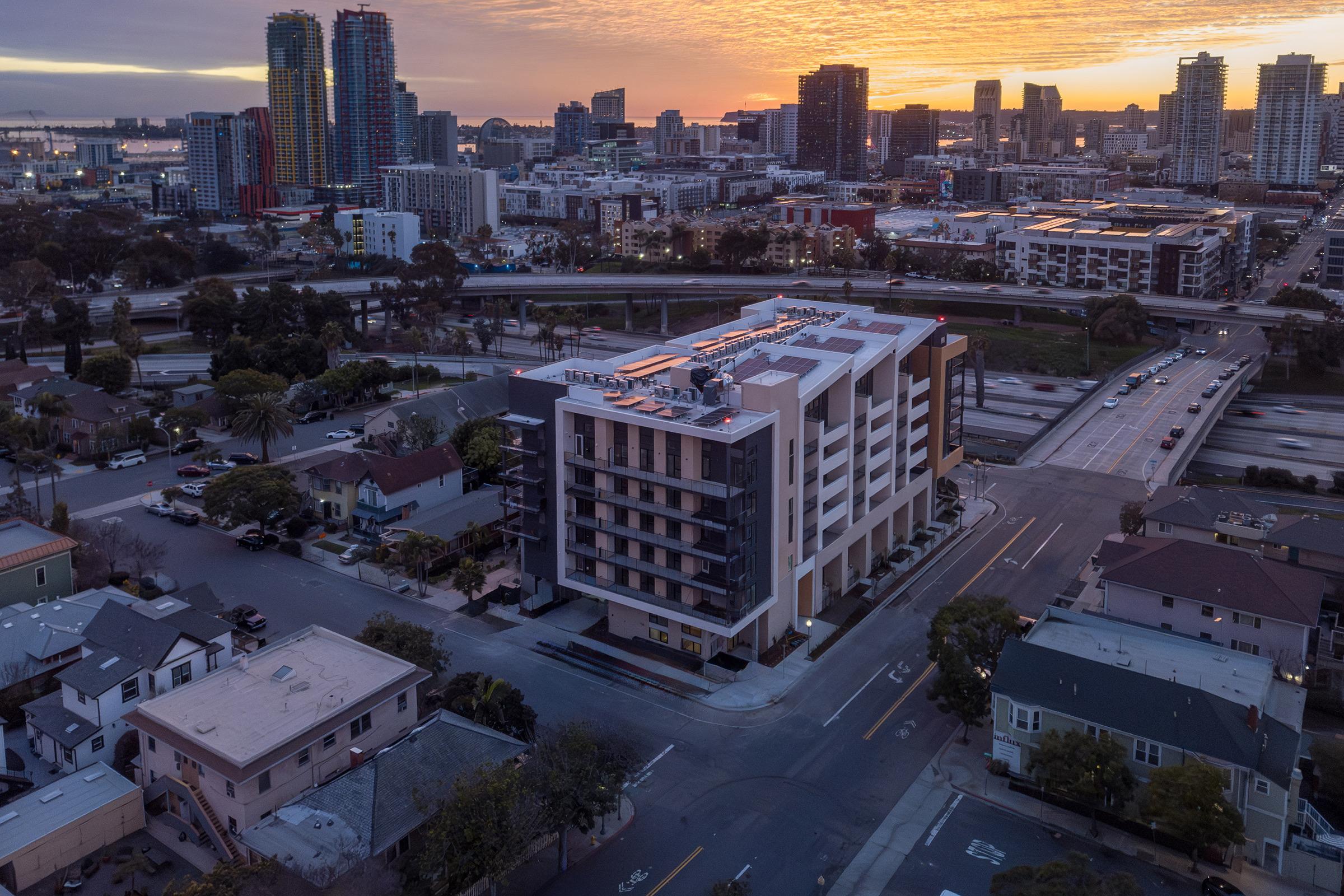 Aerial view of a modern multi-story building at sunset, surrounded by a mix of residential neighborhoods and urban development. In the background, skyscrapers and city skyline are visible, with a highway running nearby. The scene captures a blend of architecture and the vibrant colors of dusk.