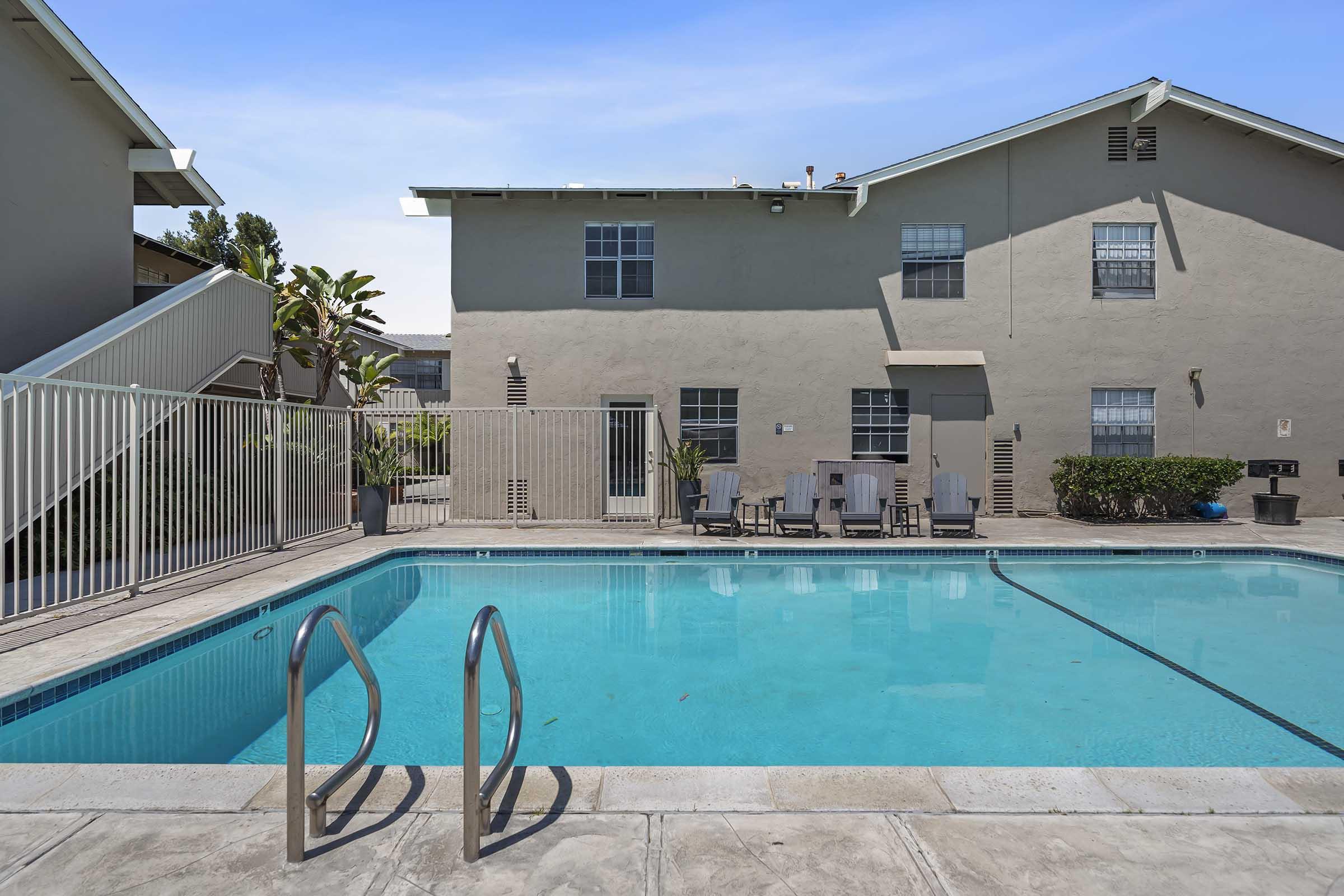 A clear swimming pool with a ladder in the foreground, surrounded by a fenced area. In the background, a two-story building with several windows and a patio area is visible. The setting is sunny with a few plants and seating arrangements near the pool.