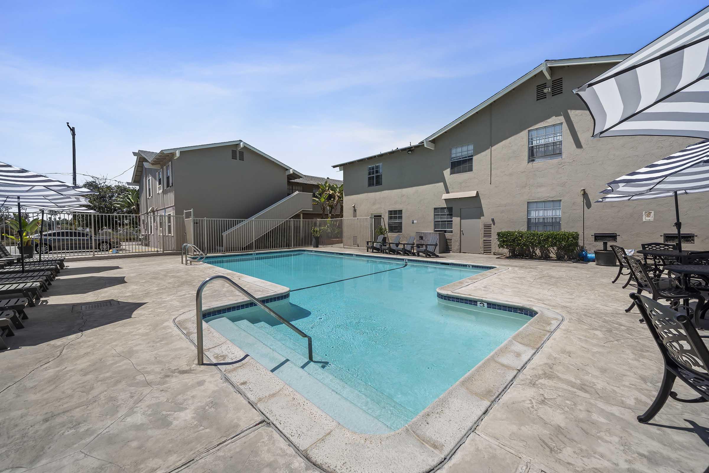 A clear swimming pool surrounded by a patio area with lounge chairs and umbrellas. In the background, there are multi-story residential buildings and lush greenery. The scene is bright and inviting, perfect for relaxation and leisure.