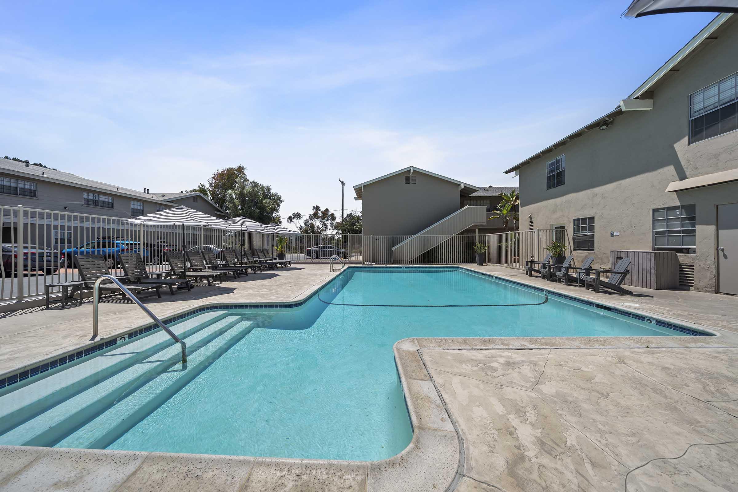 A clean and inviting swimming pool surrounded by lounge chairs and umbrellas, with two multi-story buildings in the background. The bright blue sky overhead enhances the relaxing atmosphere of the pool area, making it a perfect spot for leisure and relaxation.