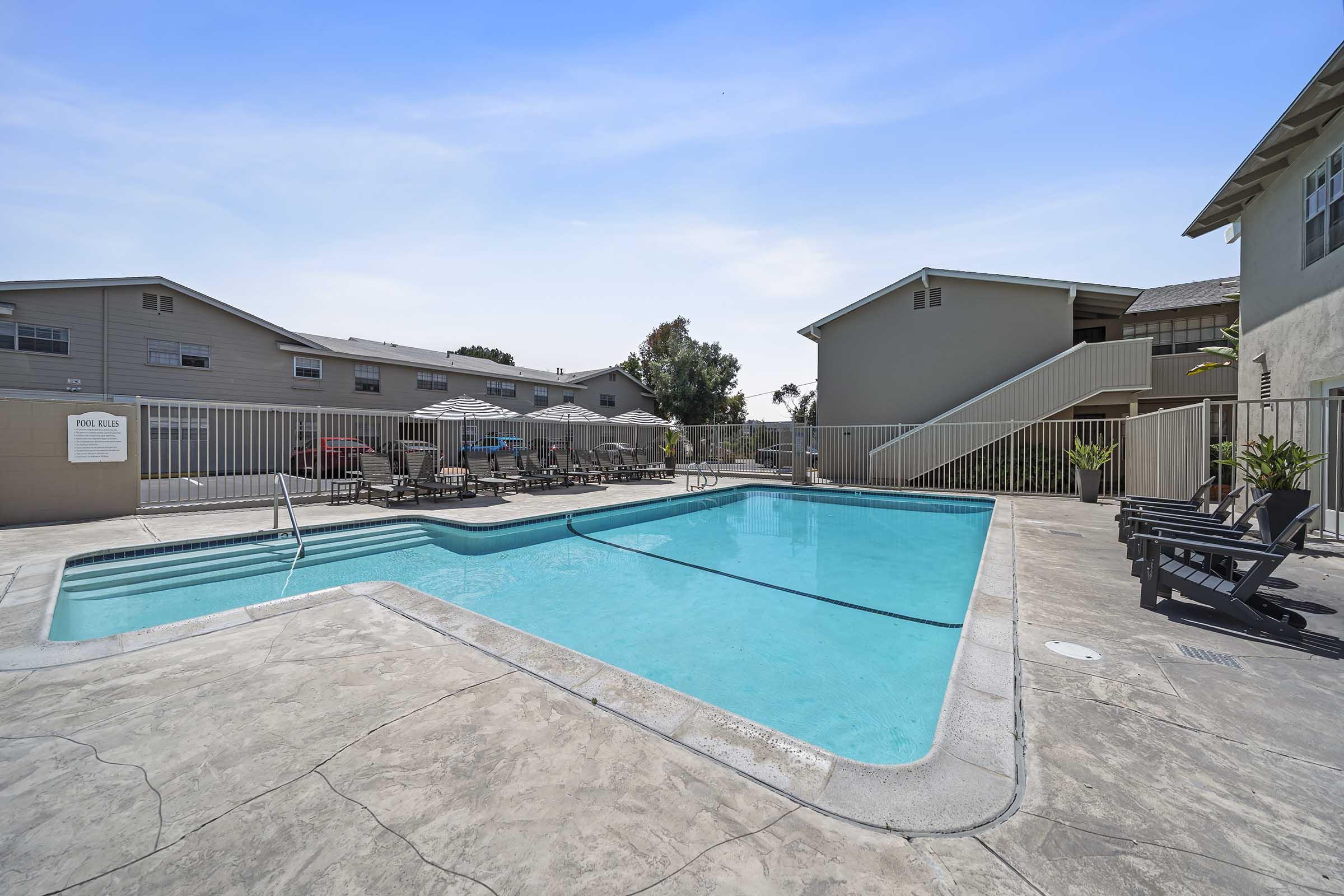 A swimming pool surrounded by a patio area with lounge chairs and umbrellas. The pool features a curved edge and is framed by a fence. In the background, there are two residential buildings with multiple stories and a clear blue sky above.