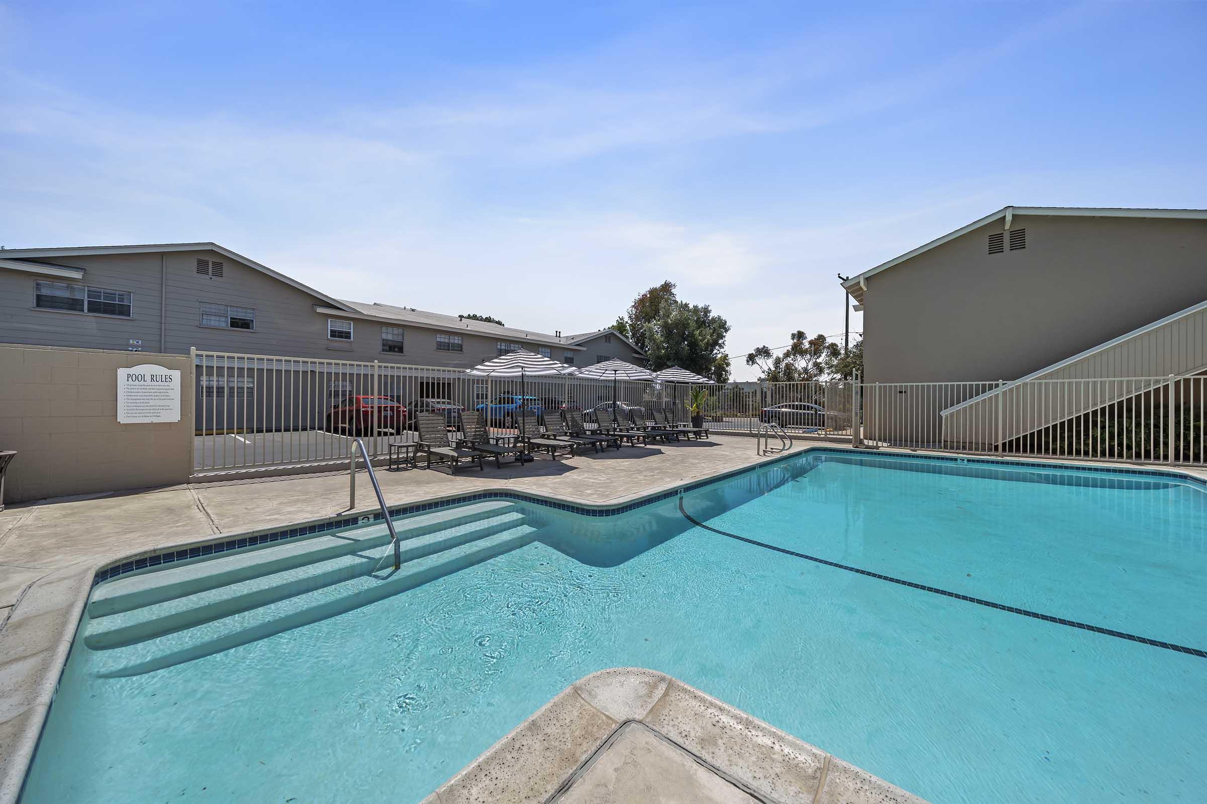 A clear swimming pool surrounded by lounge chairs and umbrellas. In the background, there are several gray apartment buildings under a blue sky. The pool area is fenced and well-maintained, offering a relaxing atmosphere.
