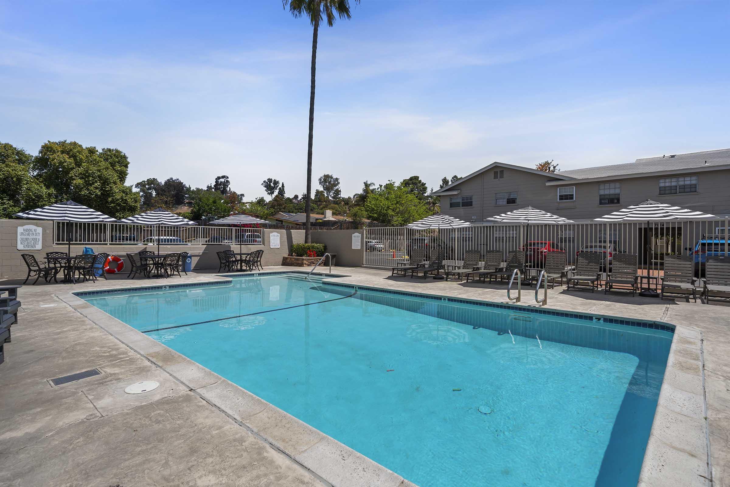 A serene outdoor pool area featuring a clear blue swimming pool surrounded by lounge chairs and patio tables. There are shaded umbrellas, a lifebuoy nearby, and a backdrop of trees and buildings, creating a relaxed atmosphere for leisure and recreation.