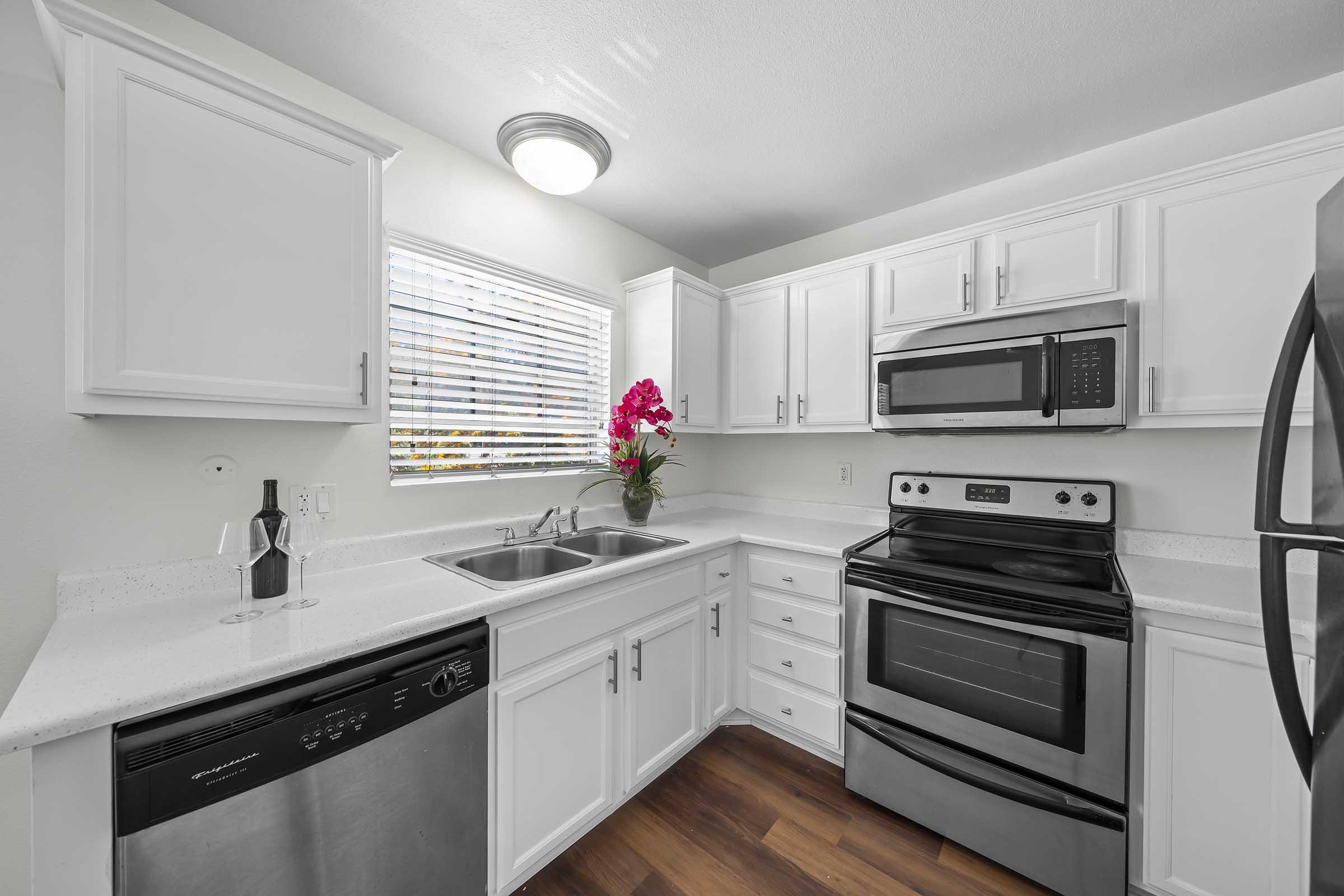 Modern kitchen featuring white cabinetry, stainless steel appliances including a microwave and stove, a dishwasher, and a sink. Natural light filters through a window with blinds, and a decorative arrangement of pink flowers adds a pop of color. The flooring is dark wood.