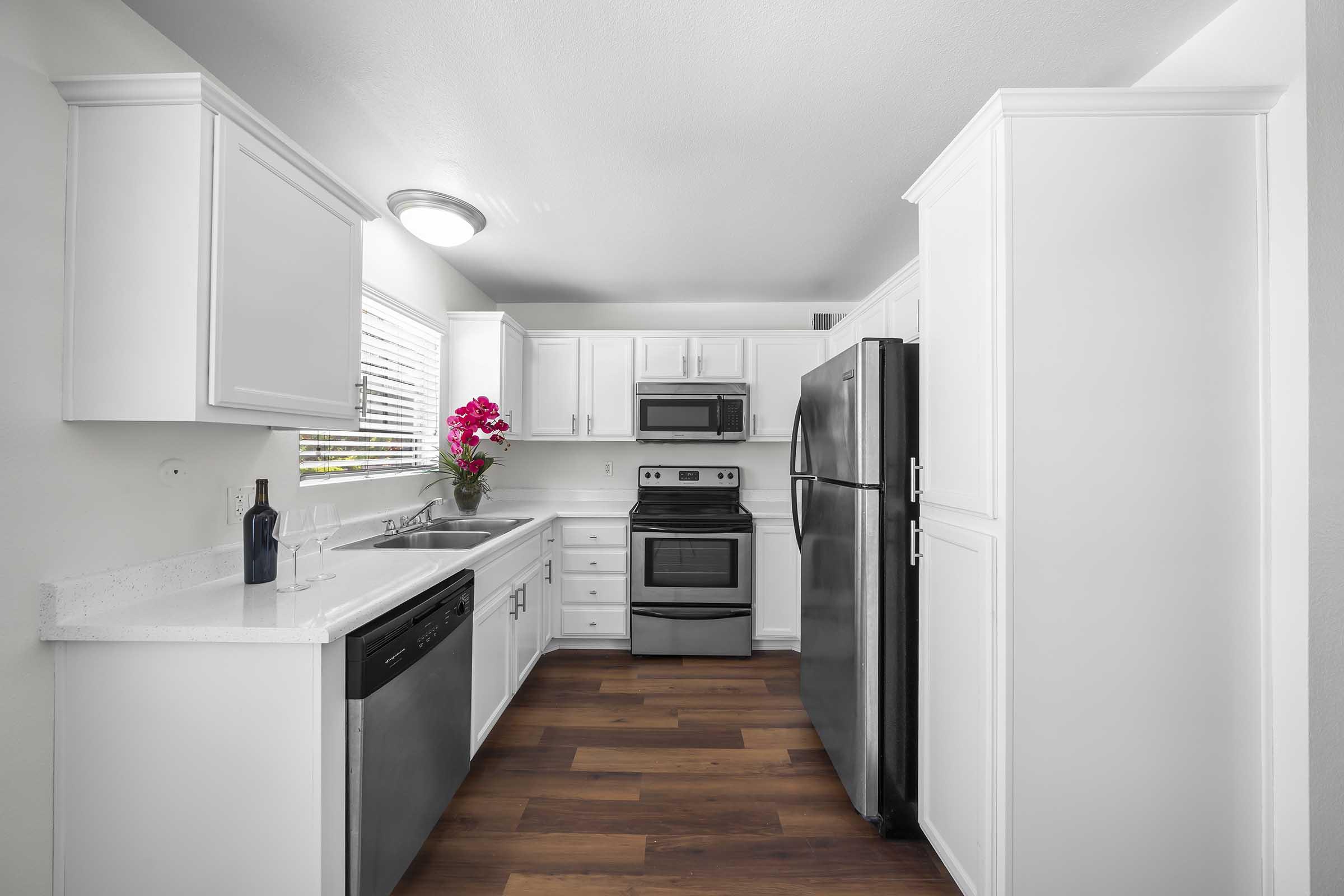 Modern kitchen featuring white cabinets, stainless steel appliances including a refrigerator and oven, a black dishwasher, and a sink. The countertop is white with a subtle pattern, and there are wooden floors. A window allows natural light in, and a potted pink flower adds a touch of color to the space.