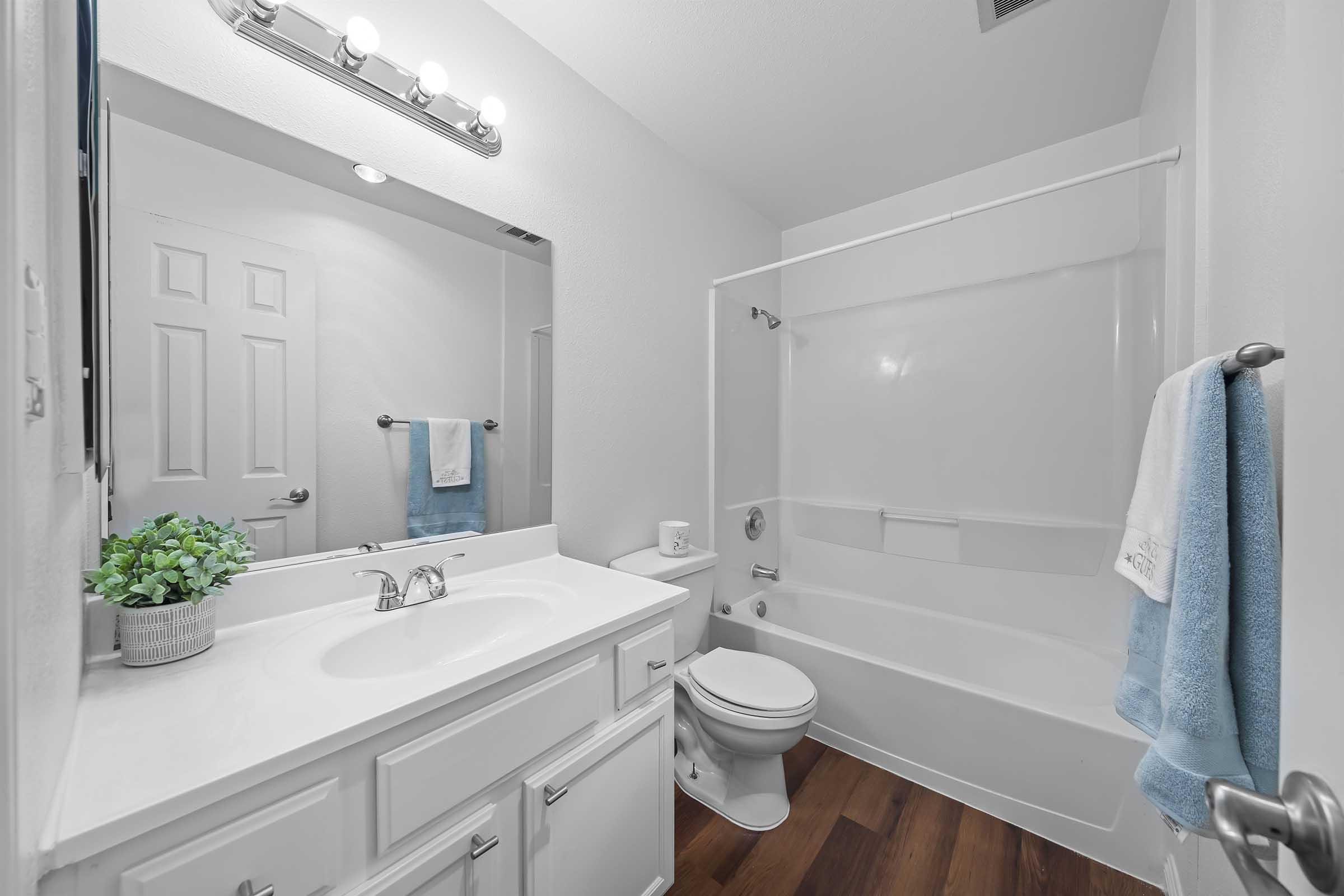 A clean and modern bathroom featuring a white vanity with a sink, a large mirror, and bright lighting. There is a bathtub with a shower curtain, a toilet, and a small potted plant on the counter. Light wood flooring adds warmth to the space, complemented by a blue towel hanging on a rack.