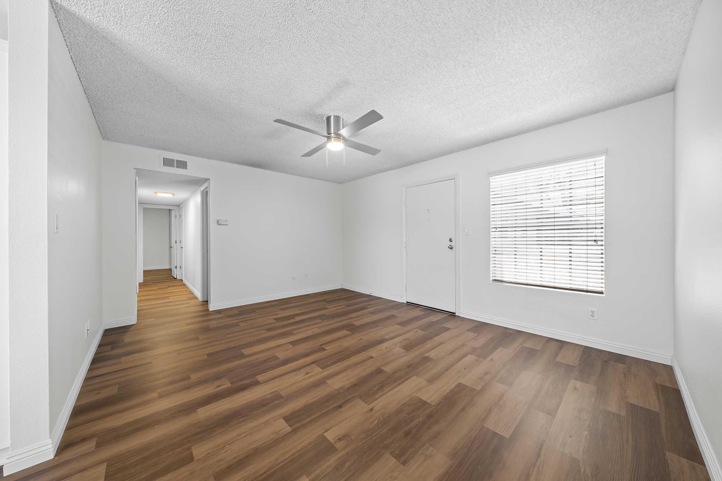 A well-lit, empty living room featuring a ceiling fan, light-colored walls, and wood laminate flooring. A paneled window with blinds is visible, along with a front door. A hallway leading to other rooms can be seen in the background. The space appears modern and inviting.