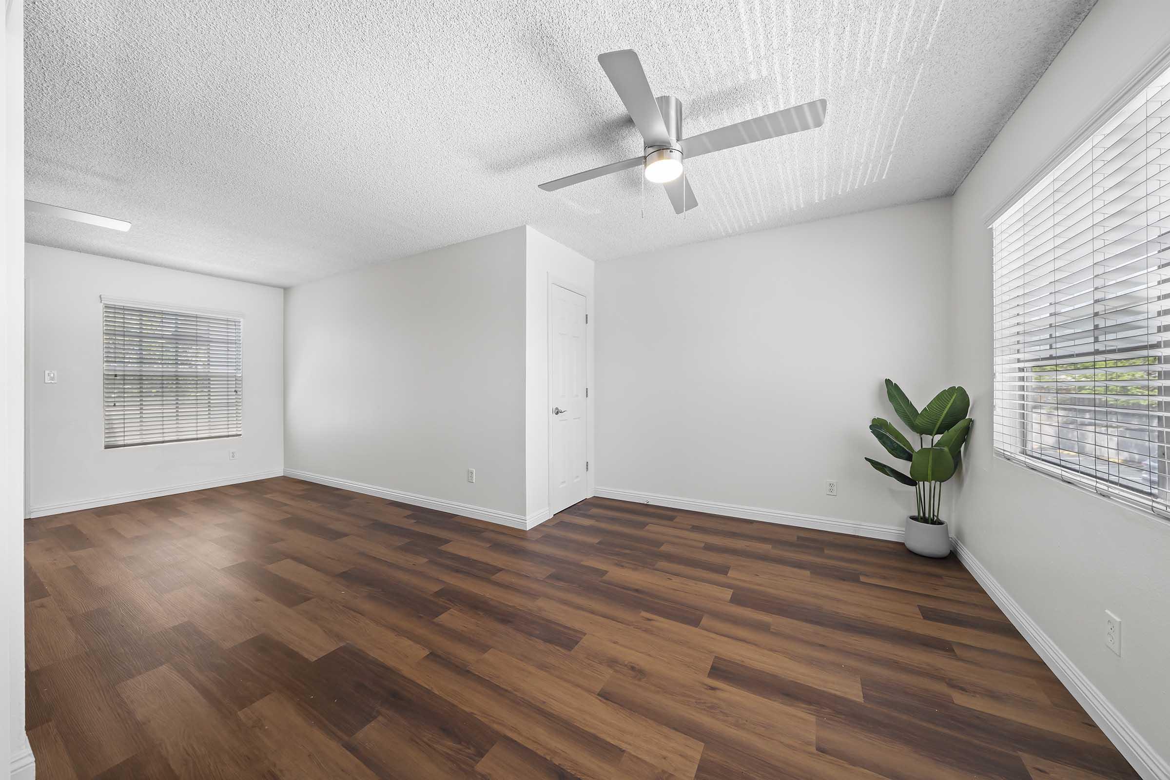 A spacious, empty room with light-colored walls and a wood laminate floor. A ceiling fan is visible, and there's a large window with blinds on one side. A potted plant sits in one corner, adding a touch of greenery to the bright, airy space.