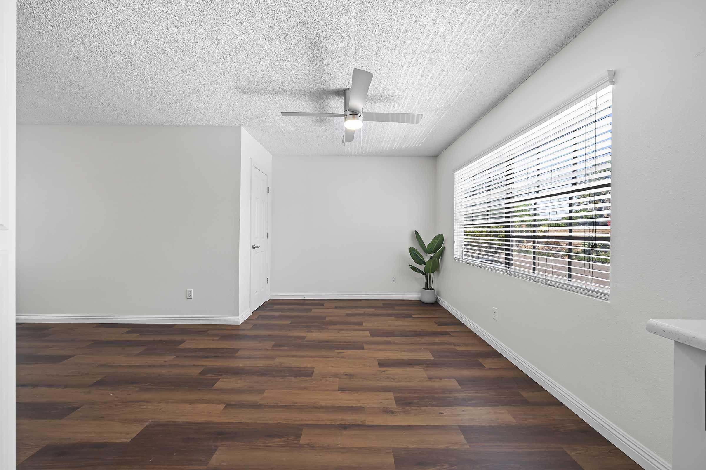 A spacious, well-lit room with a ceiling fan, featuring wooden laminate flooring. There is a potted plant near a large window with horizontal blinds, allowing natural light to enter. The walls are painted light colors, giving a fresh and airy feel to the space.