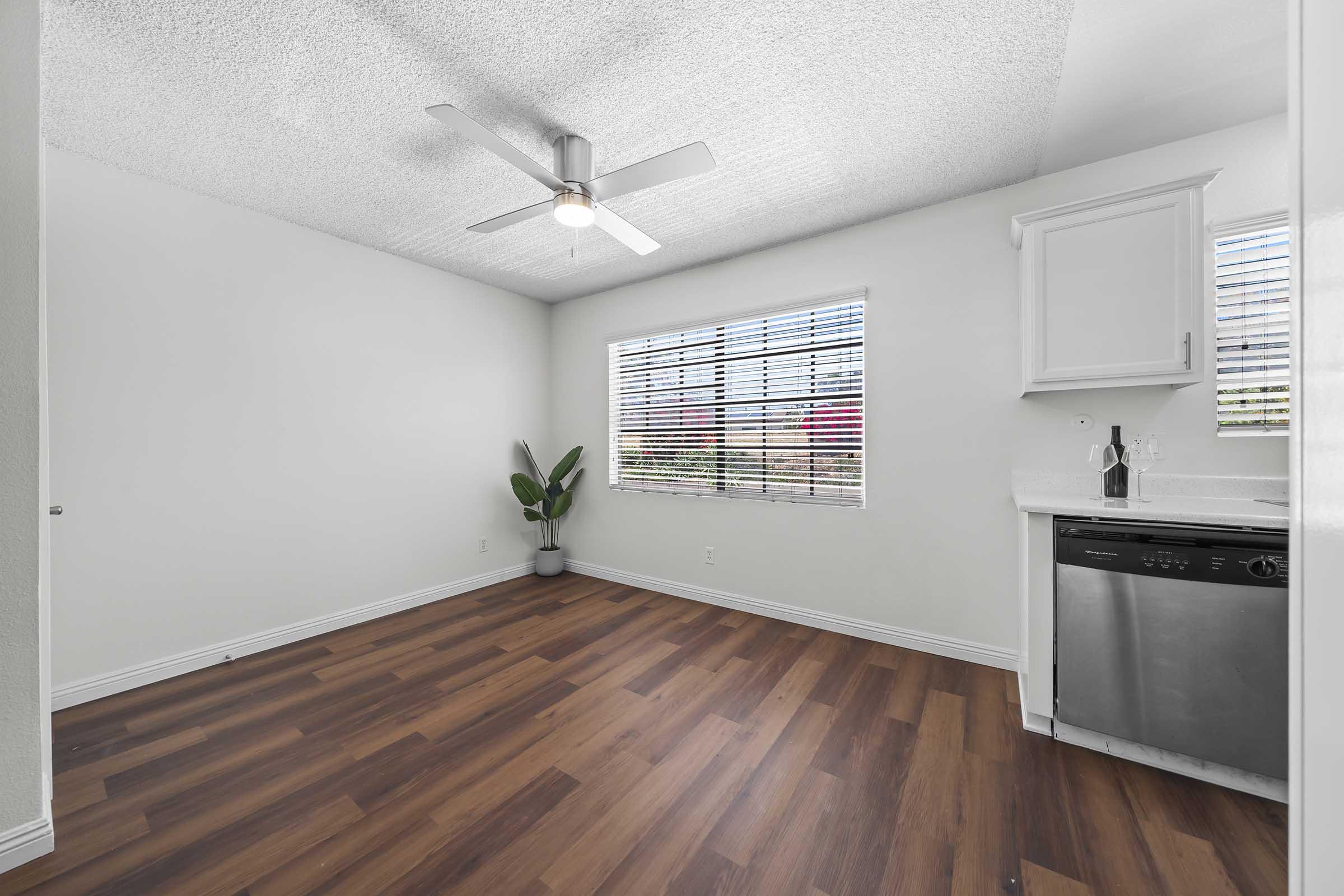 Bright and airy room featuring a ceiling fan, large window with blinds, and a small indoor plant. The space has a light-colored wall and wood-style flooring. A kitchen area is visible with a stainless steel dishwasher and white cabinetry, creating a modern and inviting atmosphere.