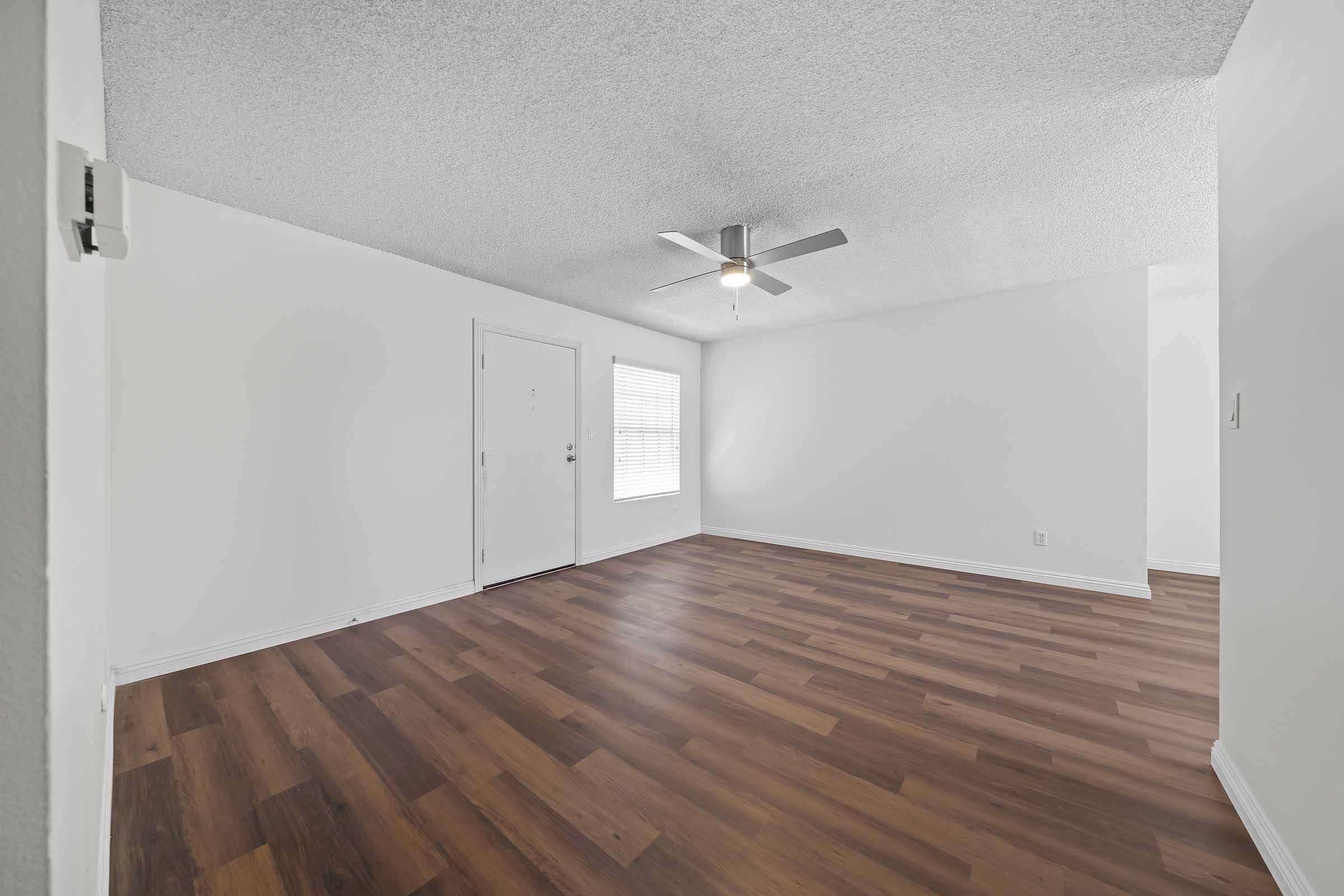 A spacious, empty room with light-colored walls and a textured ceiling. It features a ceiling fan, a window with blinds allowing natural light, and hardwood-style flooring. The entrance door is visible on the left, emphasizing the room's open and airy atmosphere.