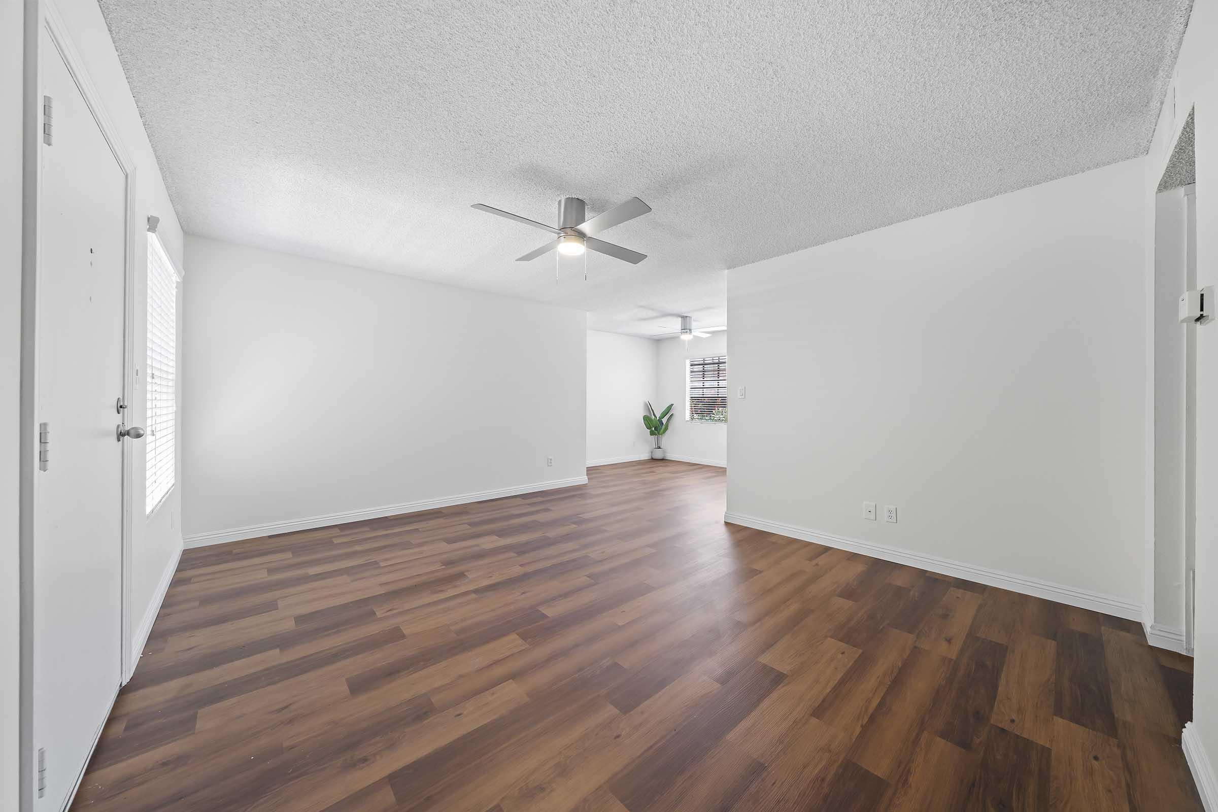 Spacious, empty room with light-colored walls and wood laminate flooring. A ceiling fan is visible, and there is a window with blinds on the left side. A small green plant sits in the corner, contributing to a bright and airy atmosphere. Ideal for various interior design options.