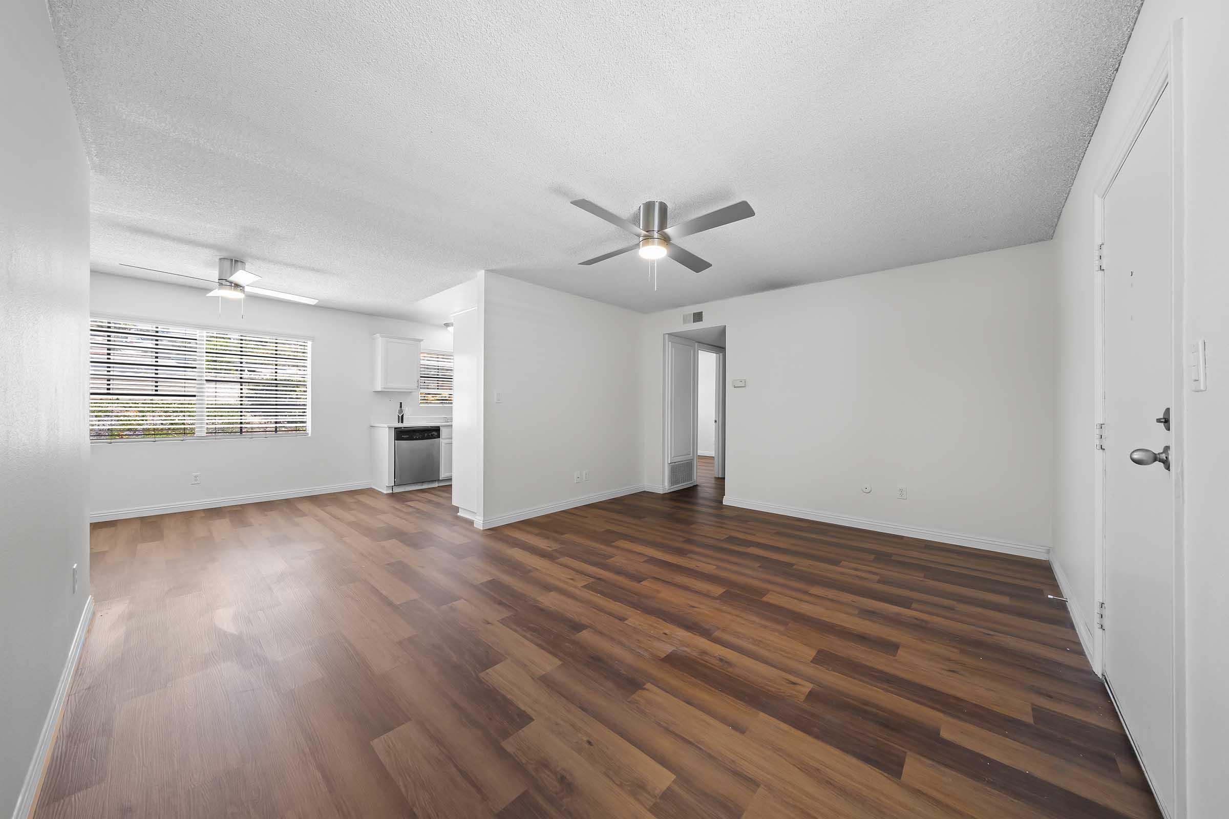 A spacious living room featuring light wood flooring, two ceiling fans, and a large window with blinds that lets in natural light. An open layout connects to a kitchen area, with a doorway leading to another room visible in the background. The walls are painted in a light color, enhancing the airy feel of the space.