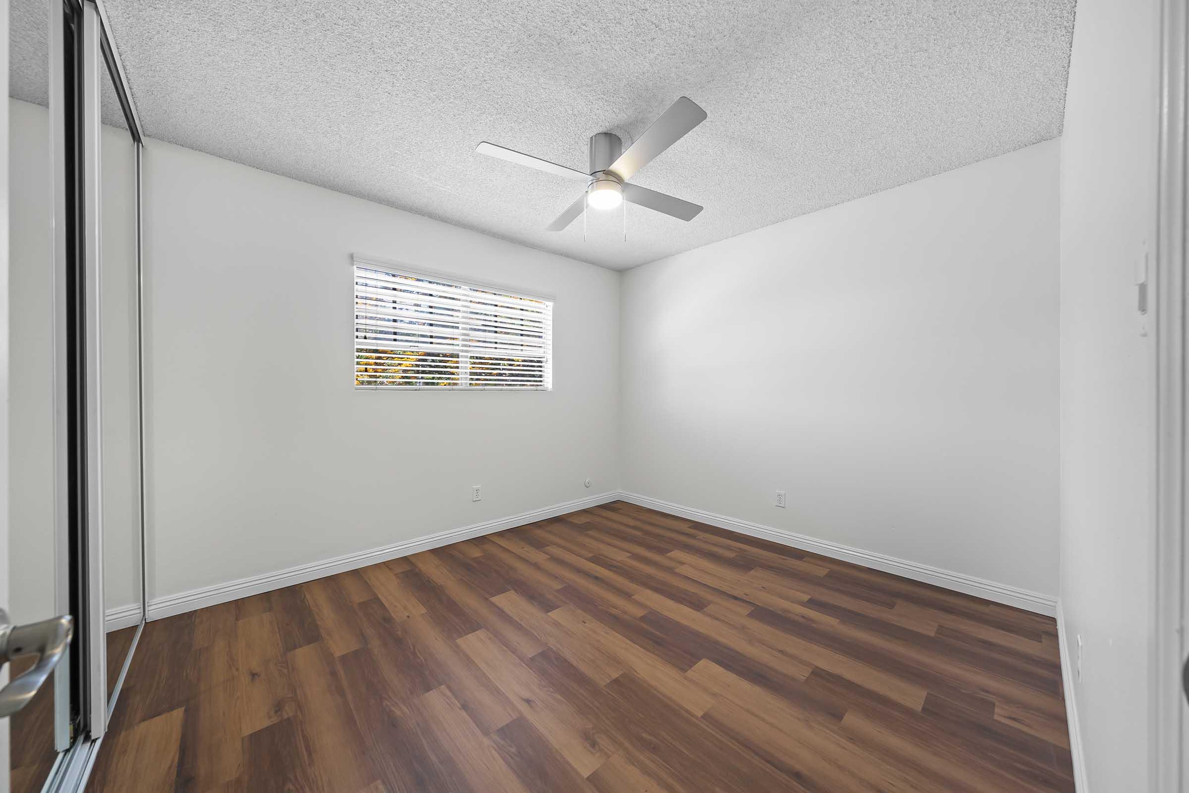 Empty room with light-colored walls and a ceiling fan. A window with blinds allows natural light to enter, and the floor features wood-like laminate. A sliding mirror closet door is visible on one side of the room.