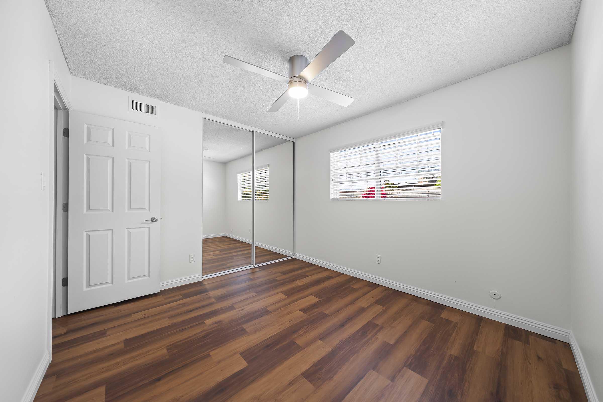 A well-lit bedroom featuring a ceiling fan, light-colored walls, and a mirrored closet door. The floor is made of brown laminate flooring, and there are two windows with white blinds, allowing natural light to enter the space. The overall ambiance is clean and modern.