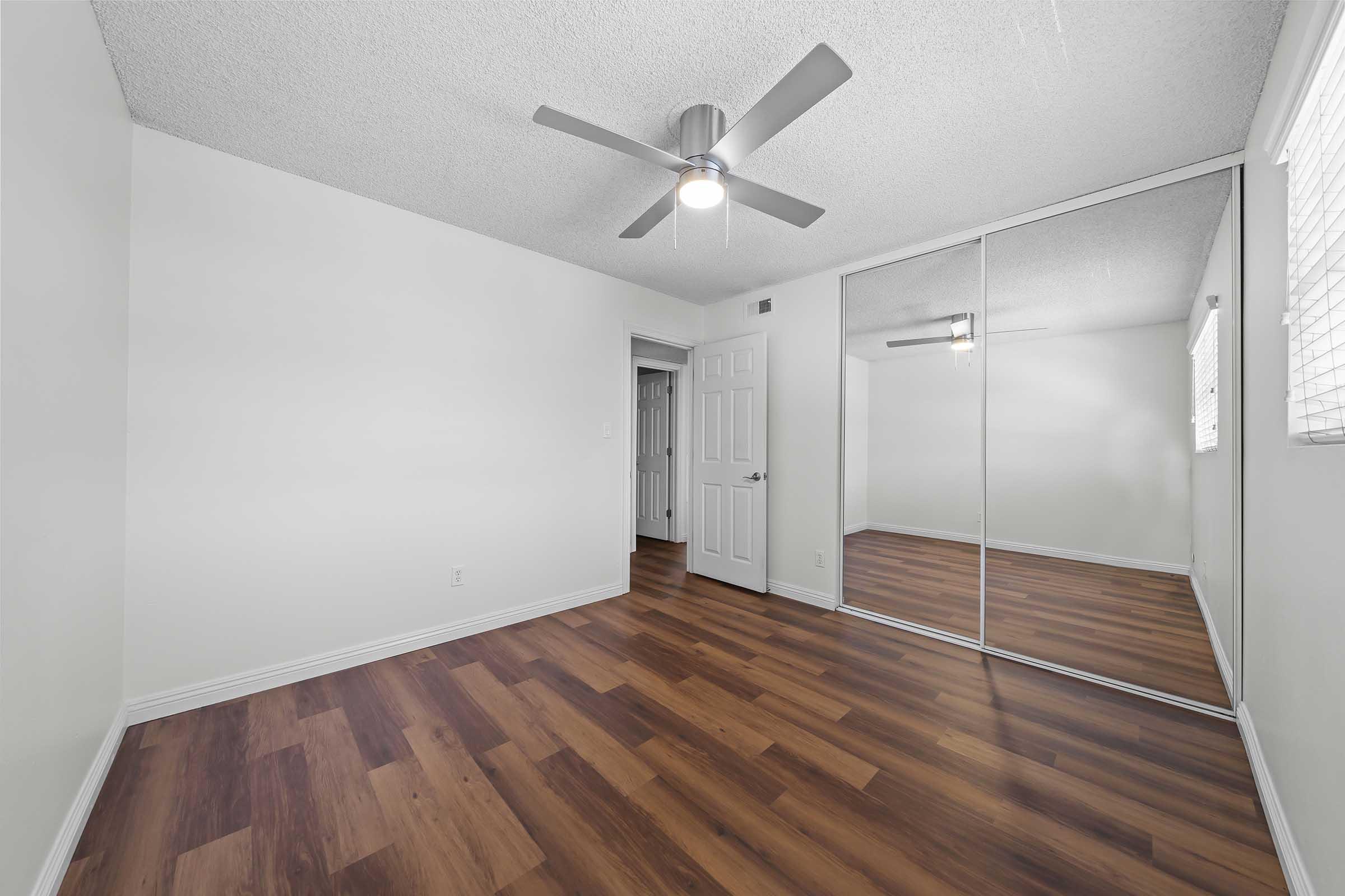 A well-lit, empty bedroom featuring light-colored walls, a ceiling fan, and wood laminate flooring. There is a mirrored closet along one wall and a door leading to another room visible in the background. Windows provide natural light, enhancing the spacious feel of the room.