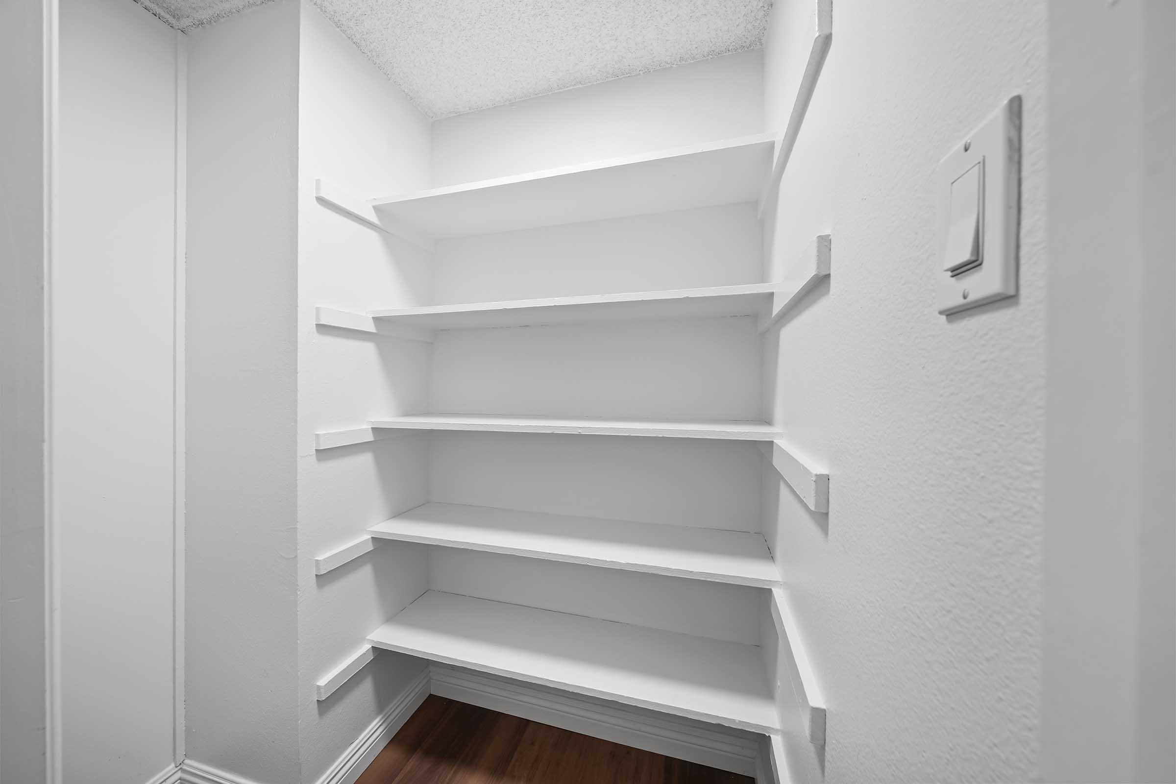 Empty white pantry with wooden shelves against a plain wall, featuring a light switch on the right side. The space is well-lit and shows no items on the shelves, emphasizing a minimalist design.