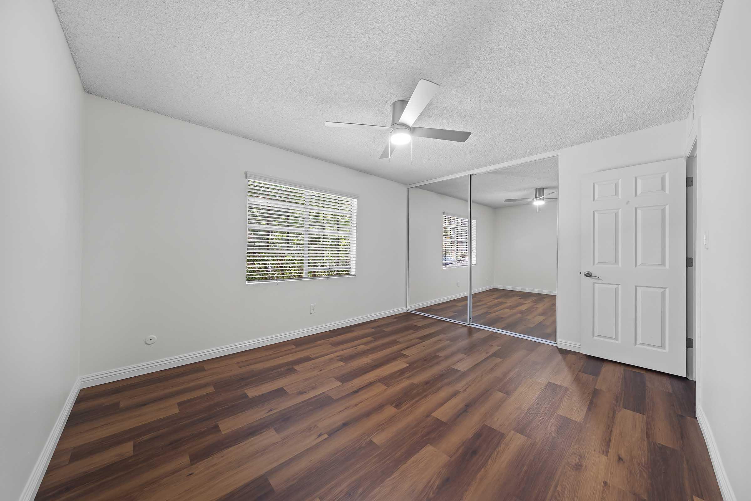 A spacious, well-lit room featuring a ceiling fan, large windows with blinds, and mirrored closet doors. The floor has a modern wood finish, and the walls are painted a soft, light color, creating an inviting atmosphere.