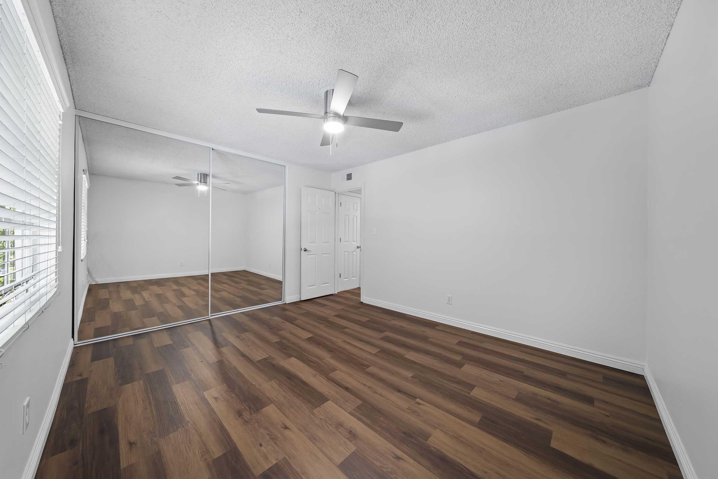 A bright, empty bedroom featuring light-colored walls and a ceiling fan. The room has mirrored closet doors reflecting the space, and the floor is covered in wooden laminate. A window with blinds allows natural light to brighten the area.
