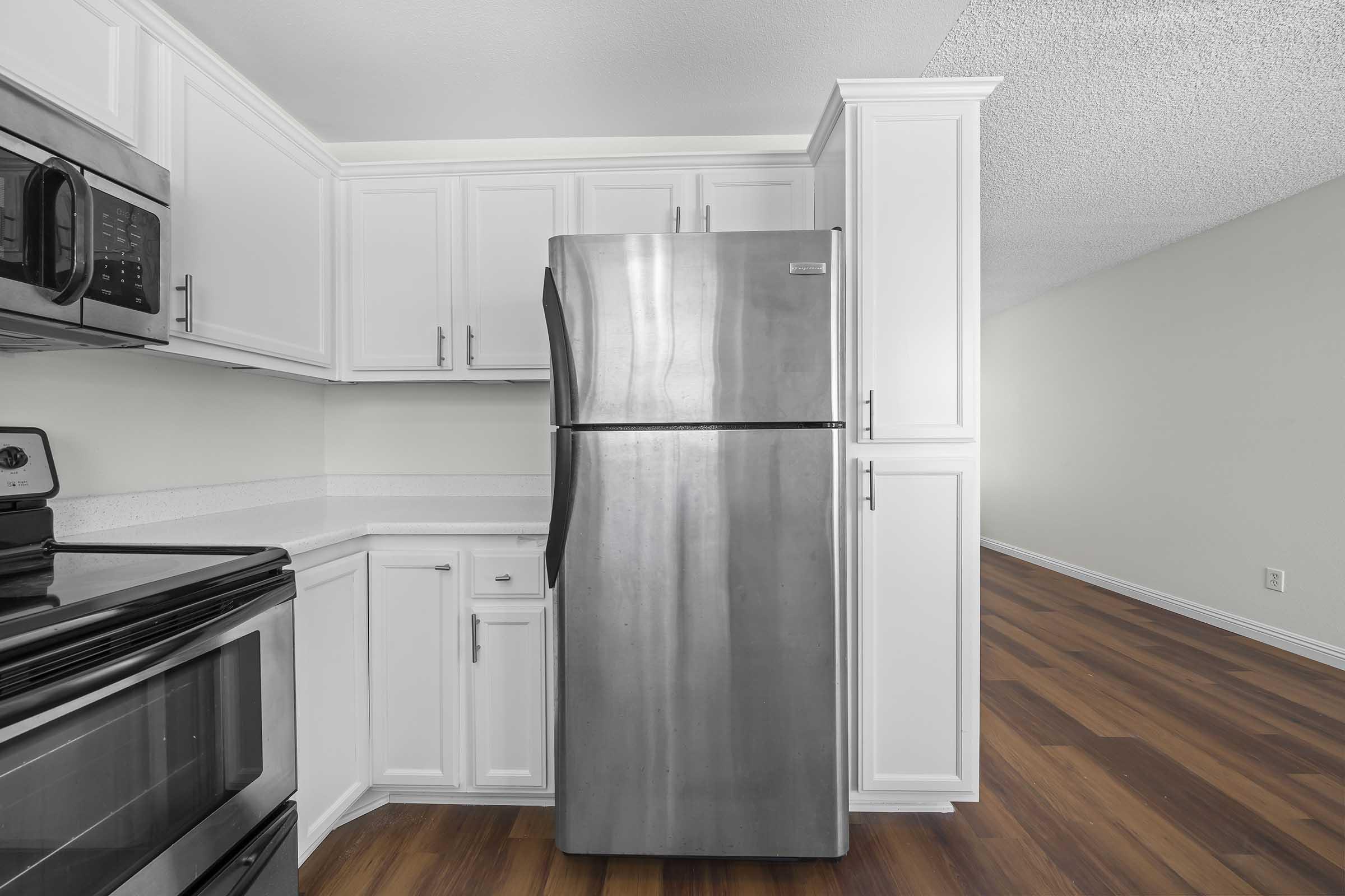 Modern kitchen featuring stainless steel refrigerator and black stove, with white cabinetry on a light green wall. The flooring is wood-like, and there's an open space suggesting a living area beyond.
